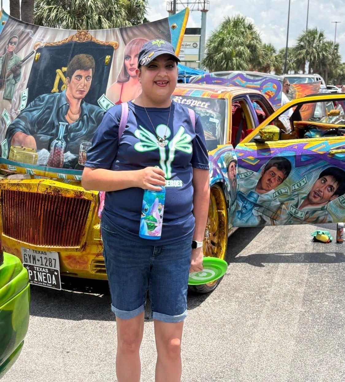 Woman stands in front of a car painted with movie scenes. She wears a blue shirt, jean shorts, and a hat.