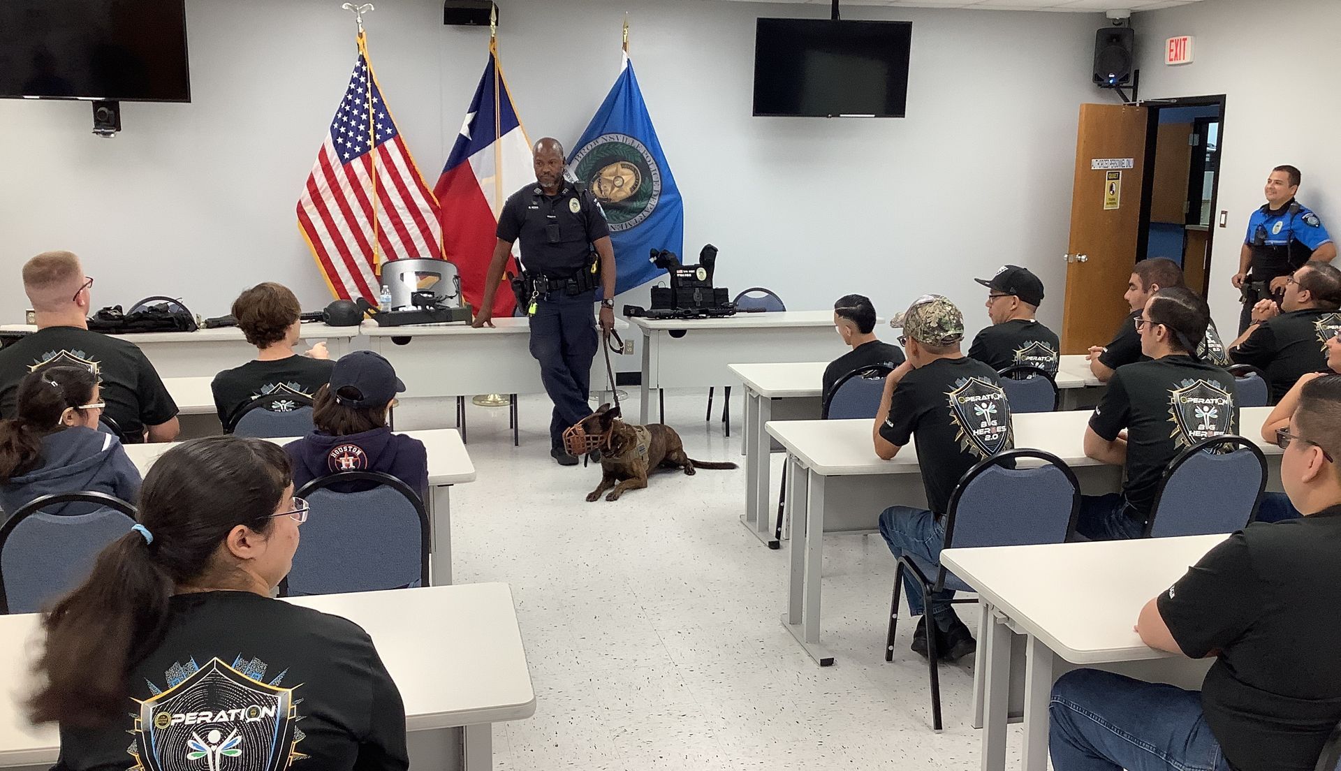 A group of people are sitting at tables in a classroom with a police officer standing in front of them.