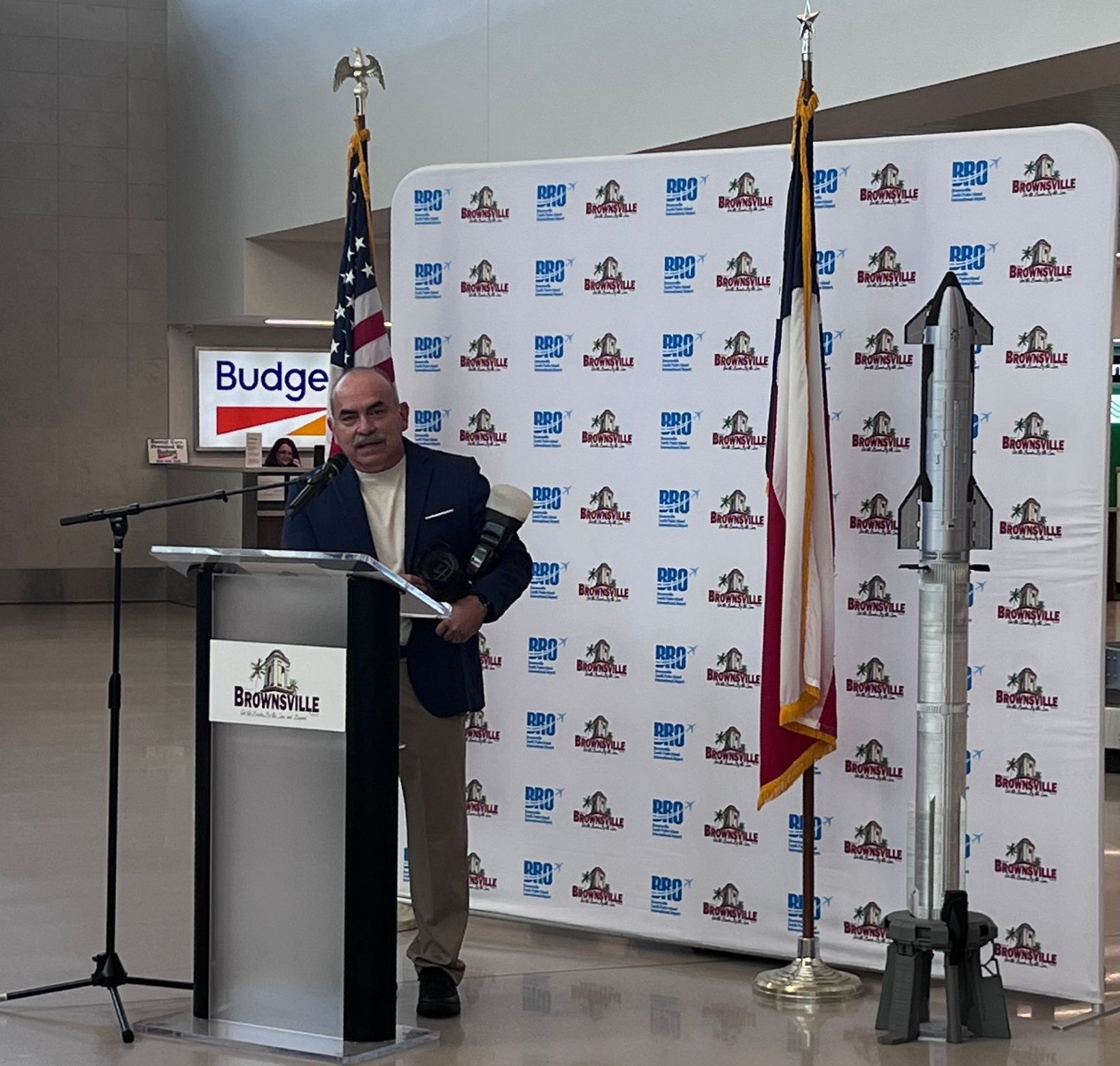 A man stands at a podium in front of a wall that says budget