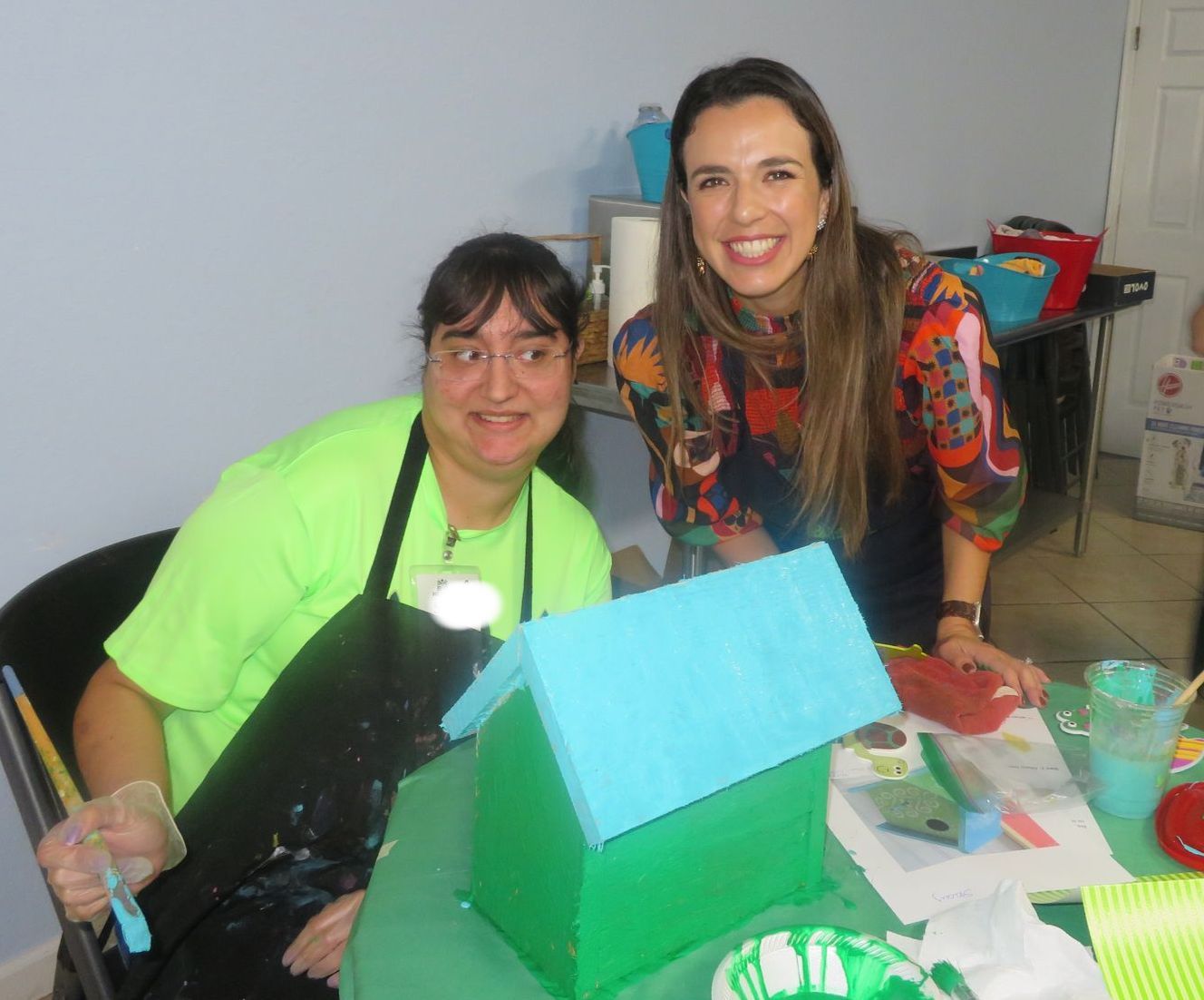 Two women are sitting at a table with a model house in front of them