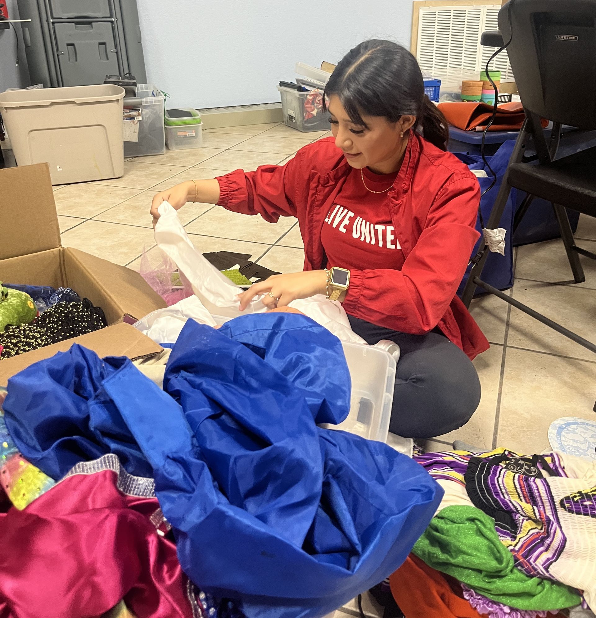 A woman wearing a red shirt that says live onate is kneeling down in front of a pile of clothes.