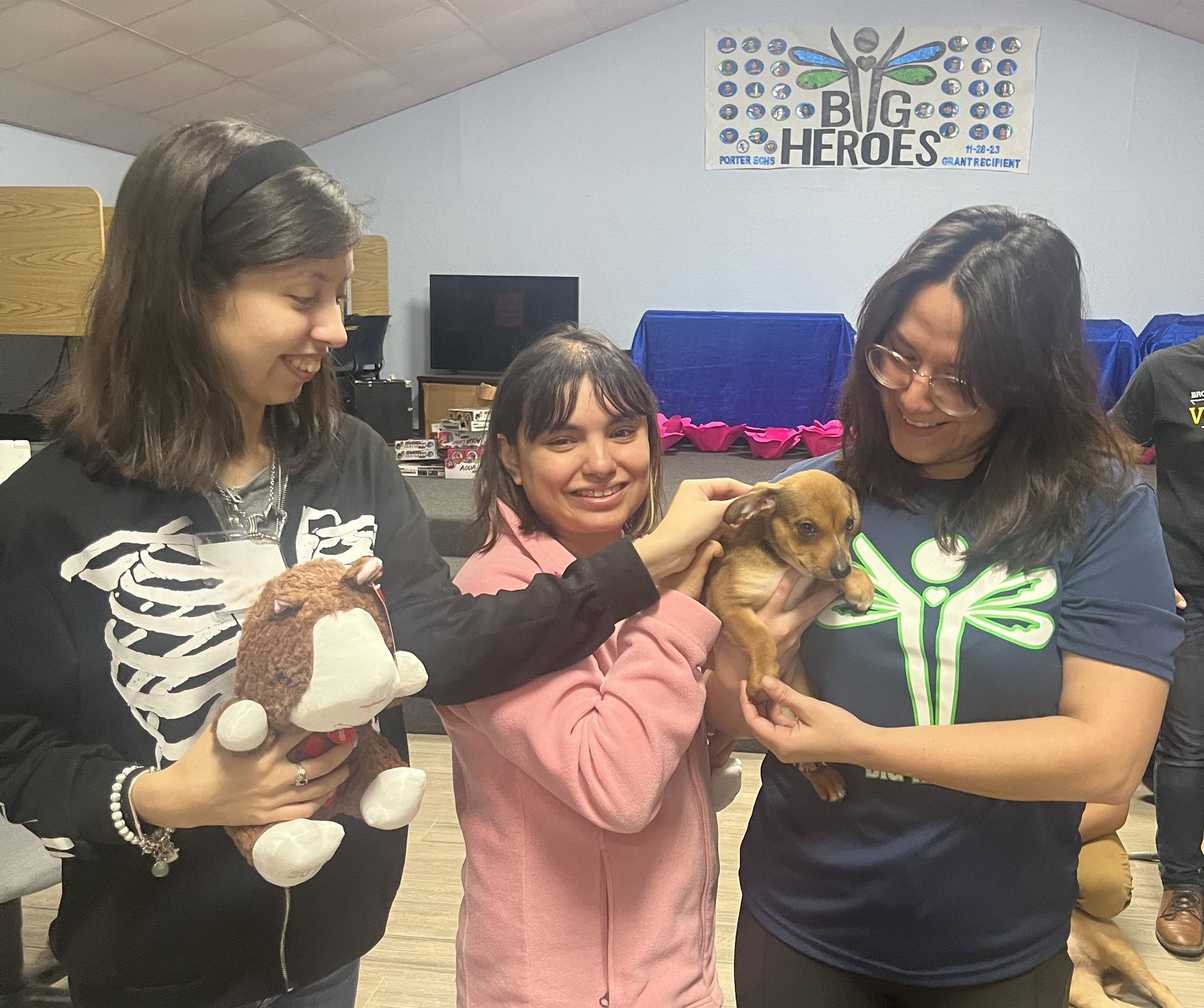 Three women are holding a puppy in front of a sign that says big heroes