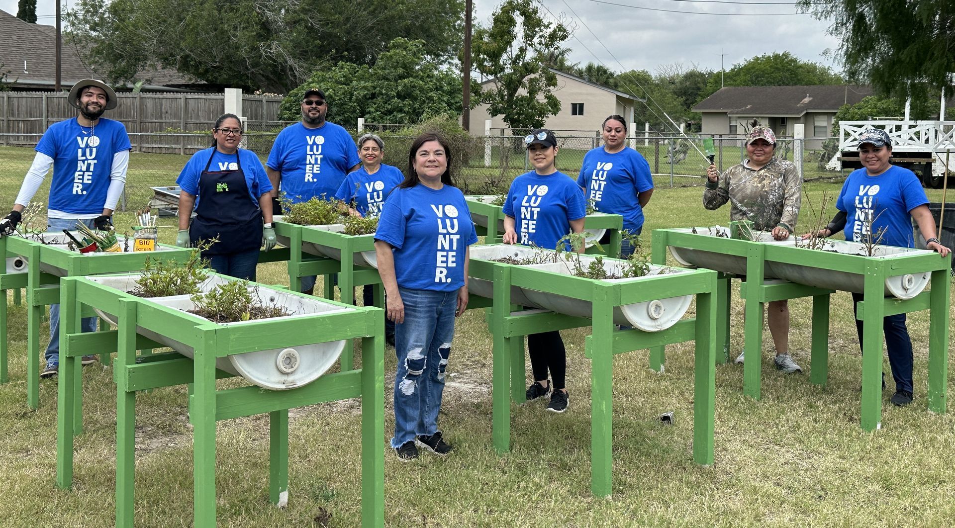 A group of people are standing in front of green tables in a field.