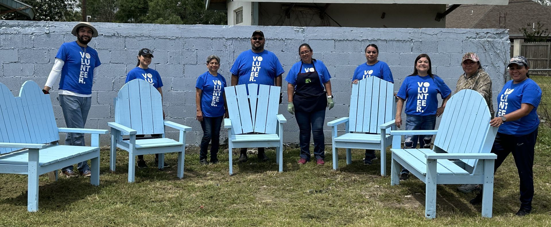 A group of people are standing in front of a wall holding blue chairs.