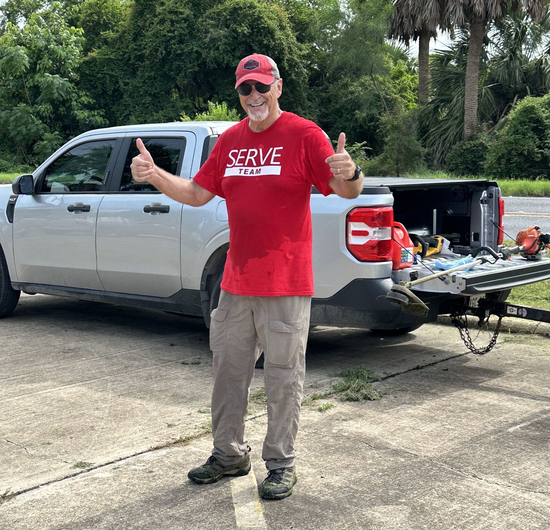 A man in a red shirt is giving a thumbs up in front of a truck.