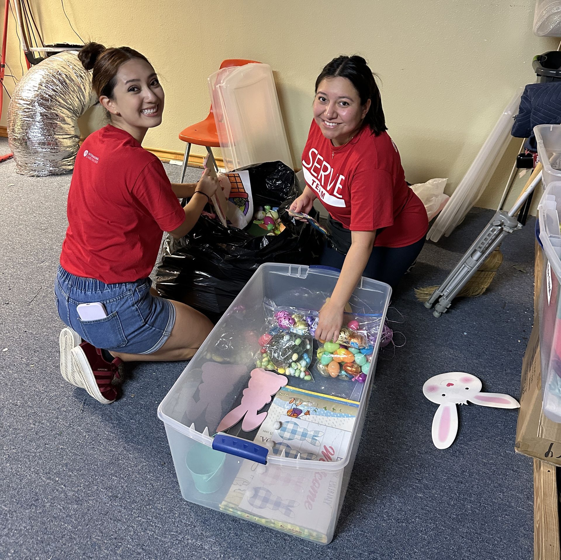 Two women in red shirts are kneeling next to a plastic bin filled with toys.