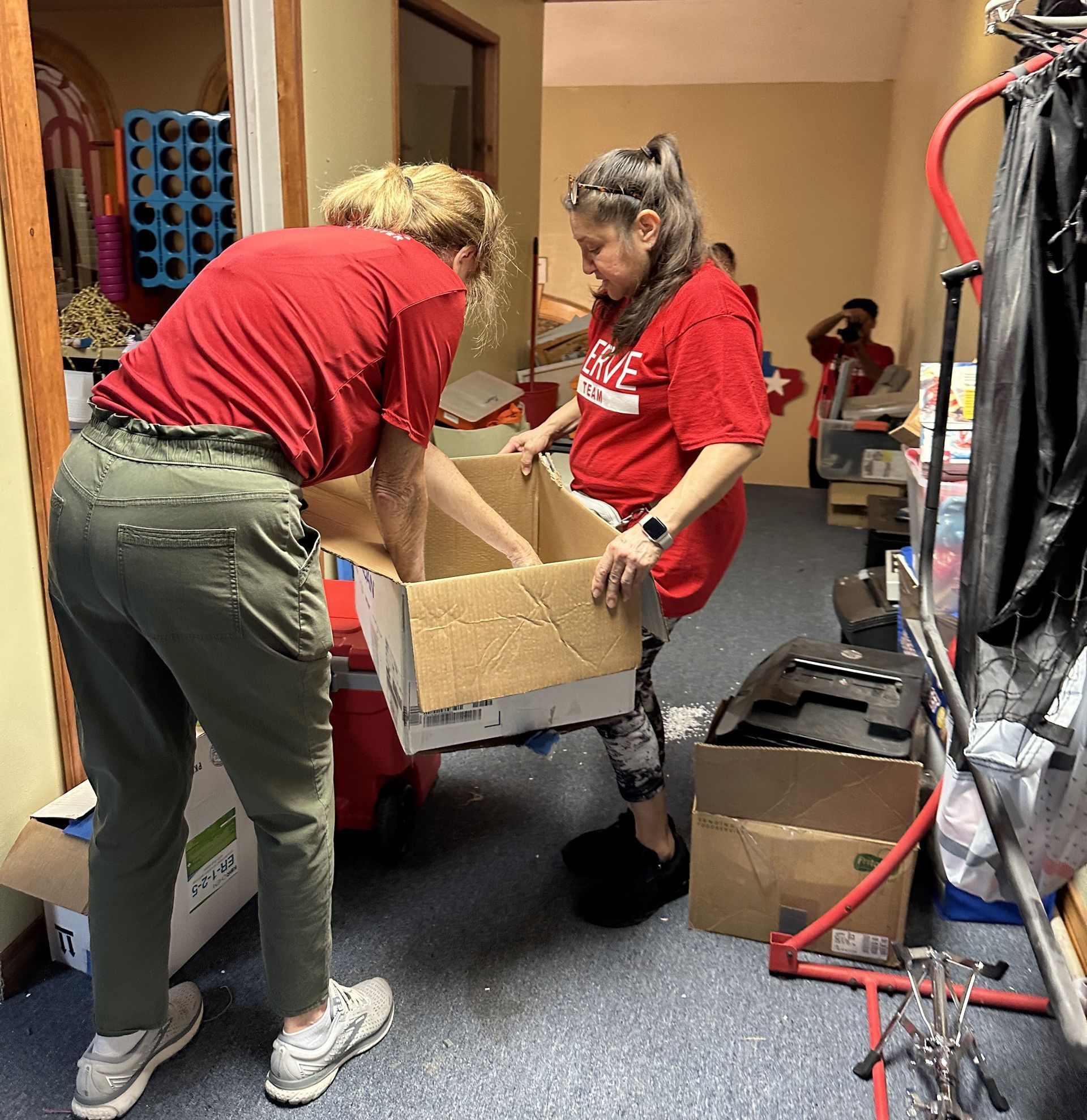 Two women in red shirts are pushing a cardboard box