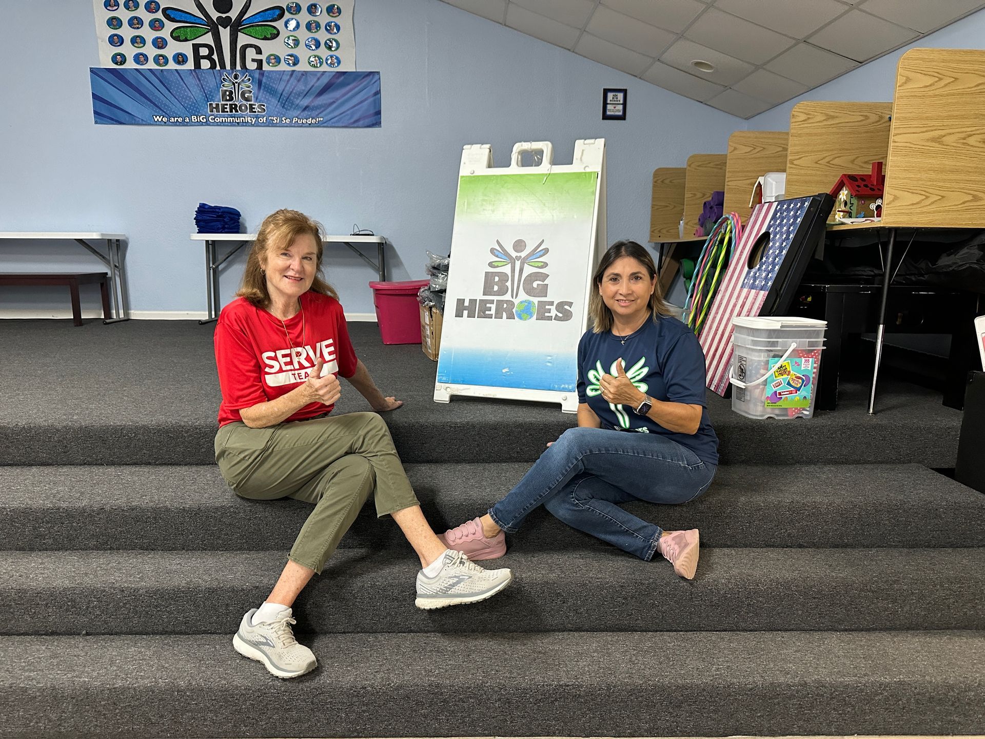 Two women are sitting on a set of stairs next to a sign that says big heroes.
