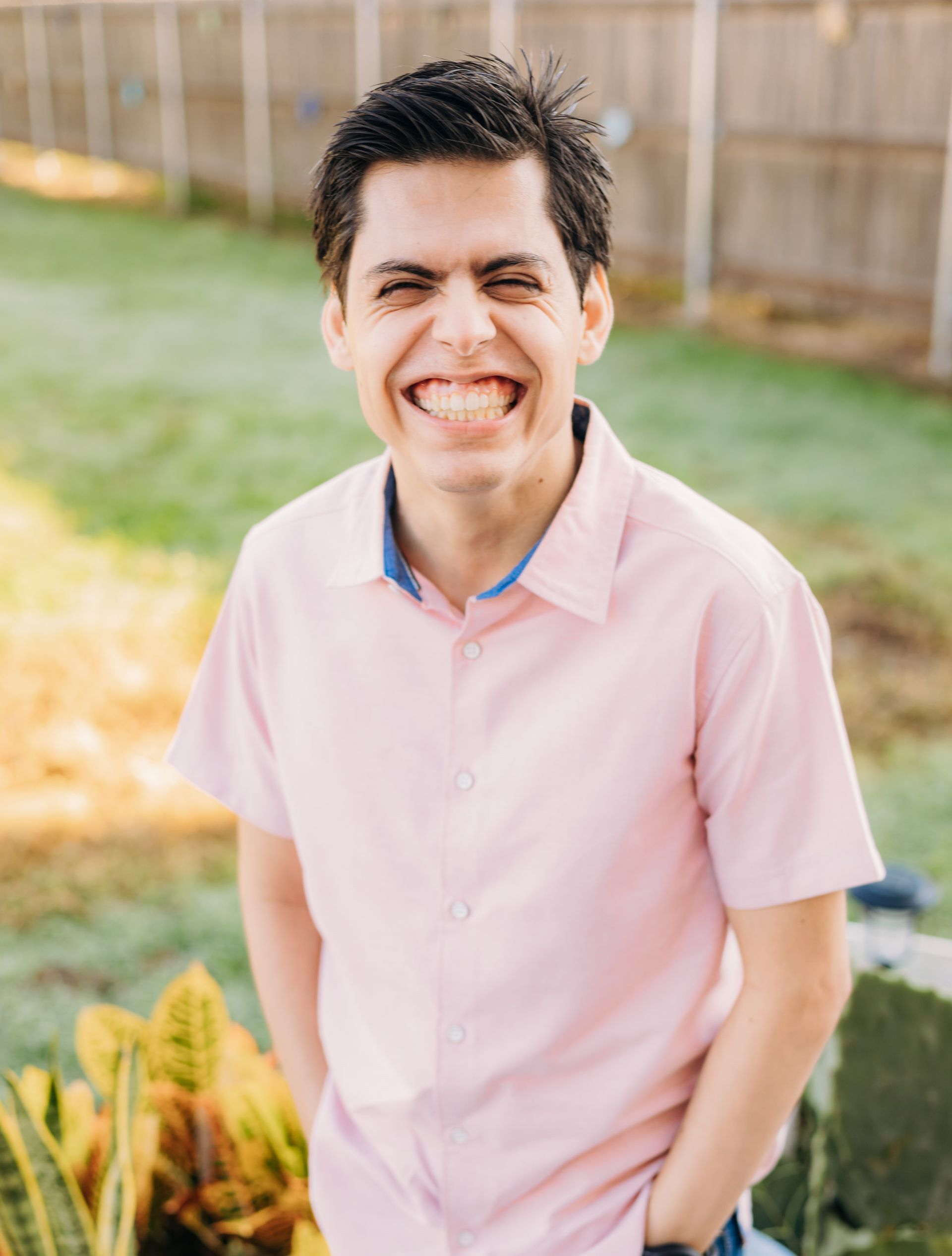 A man in a pink shirt is standing in front of a fence and smiling.