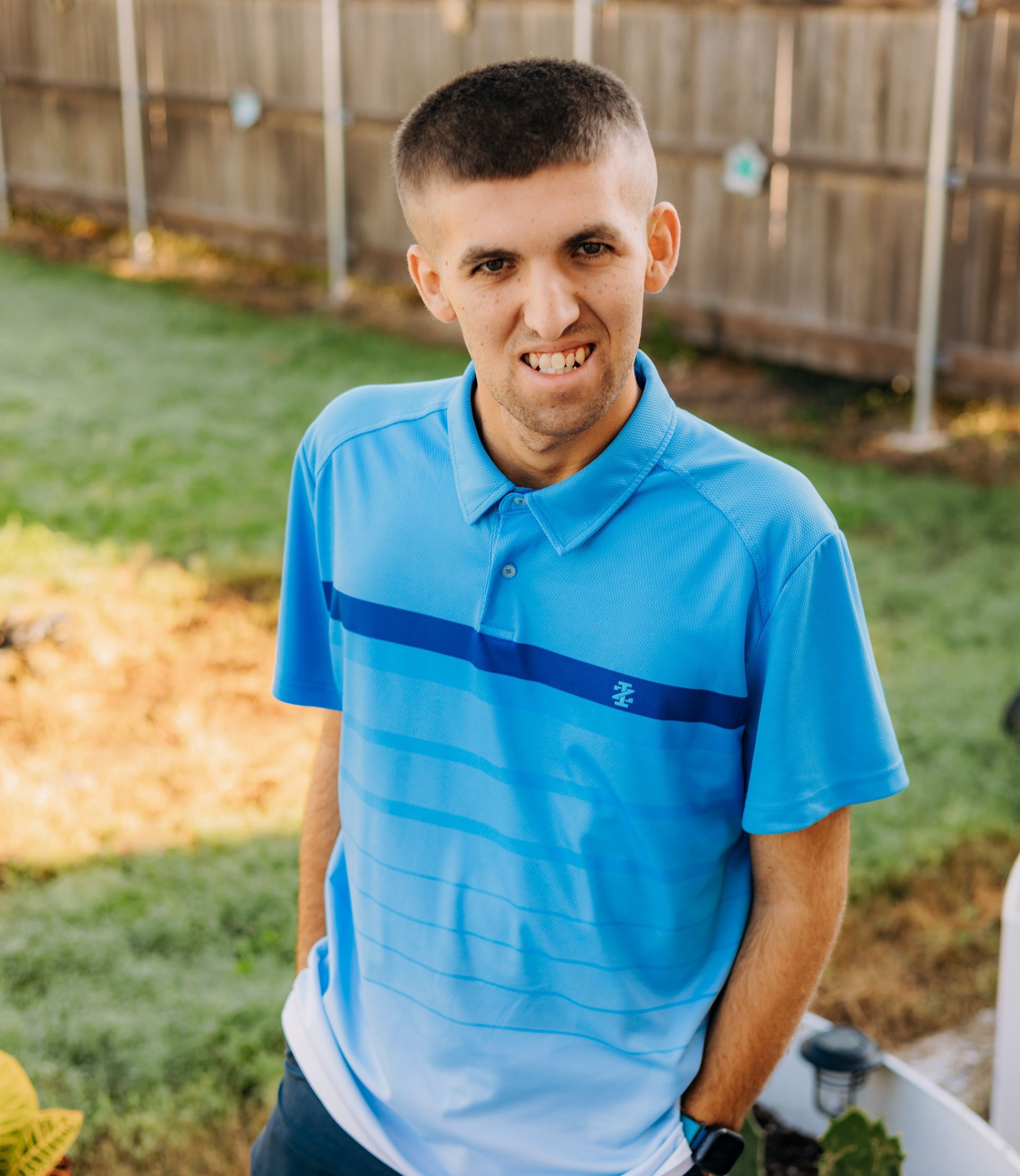 A man in a blue polo shirt is standing in a backyard with his hands in his pockets.