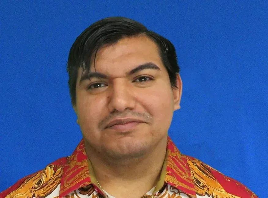 A man in a red shirt is standing in front of a blue background.