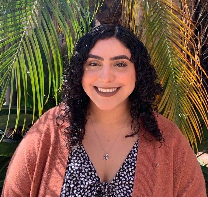 A woman with curly hair is smiling in front of a palm tree.