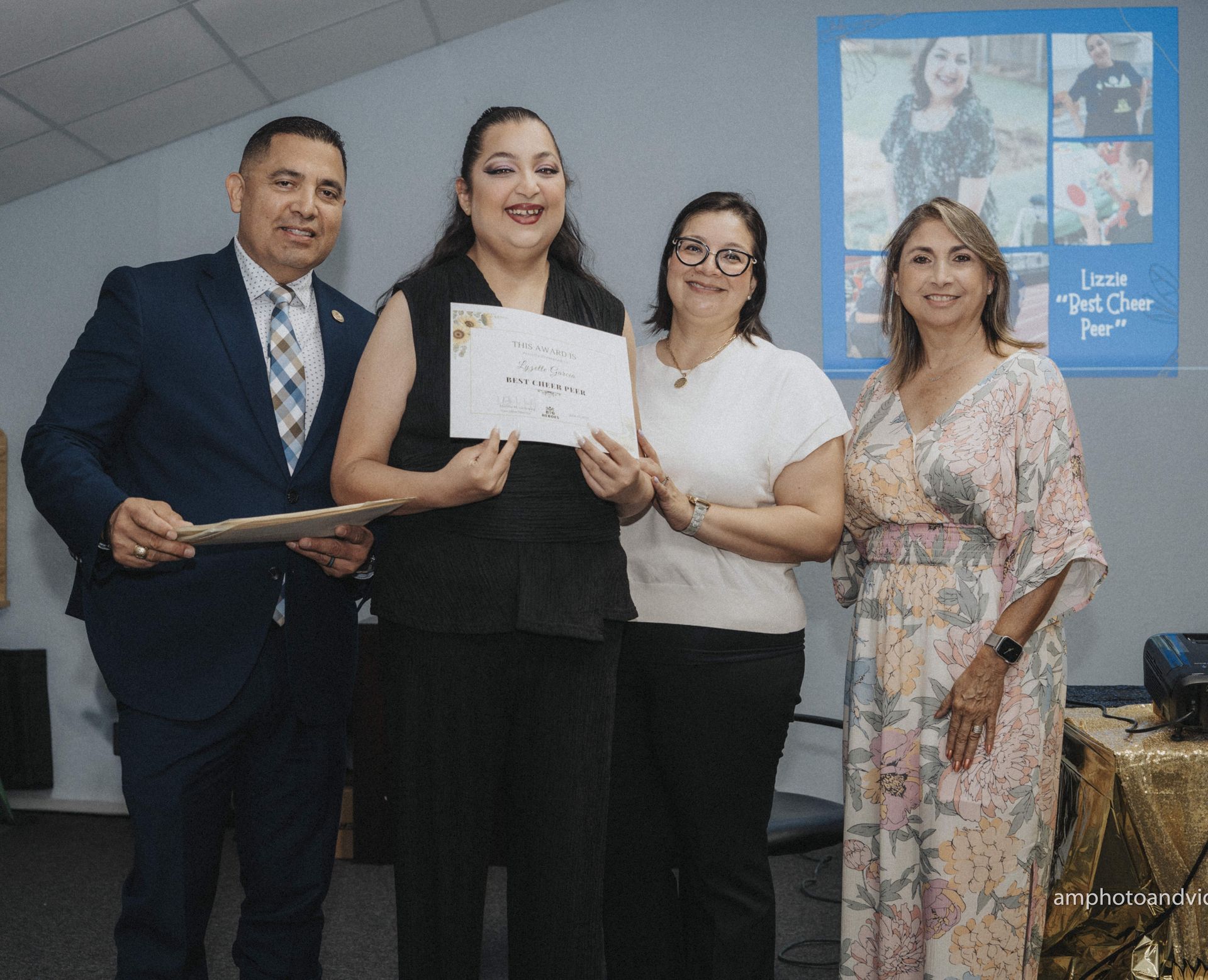 Group of four people at an event, holding a certificate. Smiling, formal attire, a blue background.
