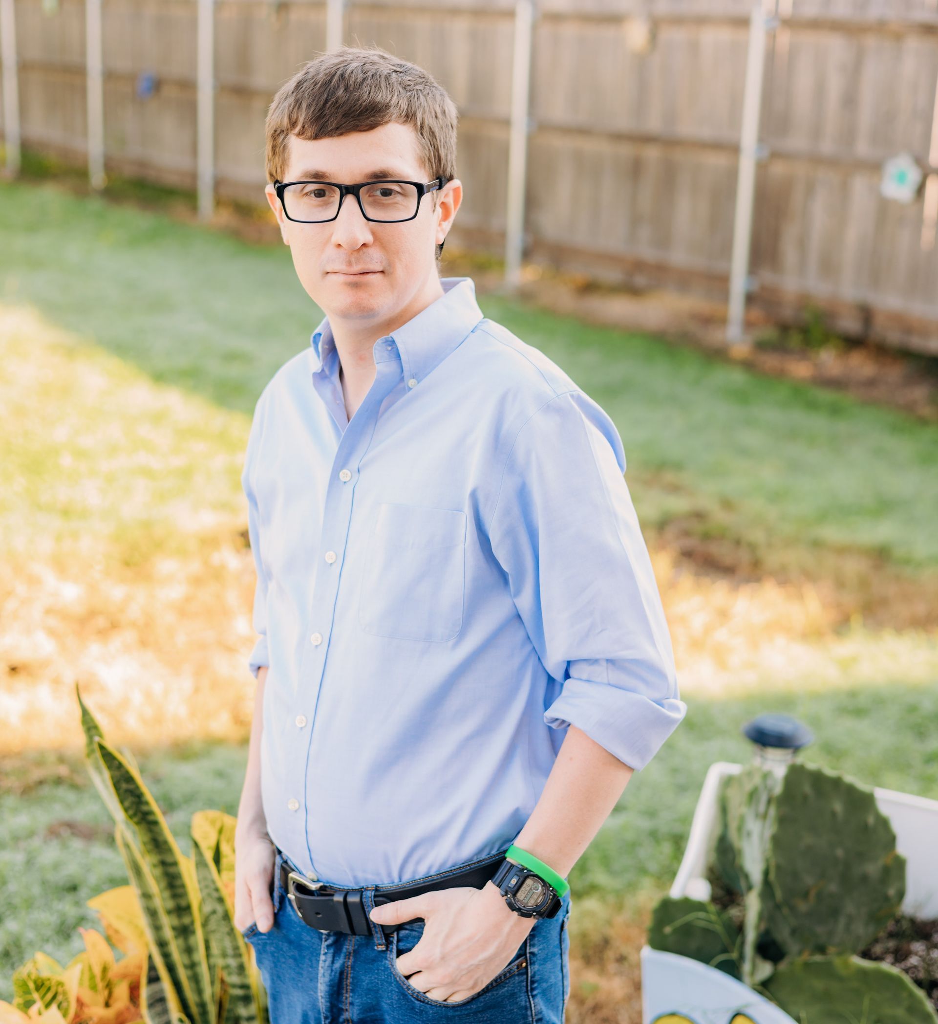 A man wearing glasses is standing in a garden with his hands in his pockets