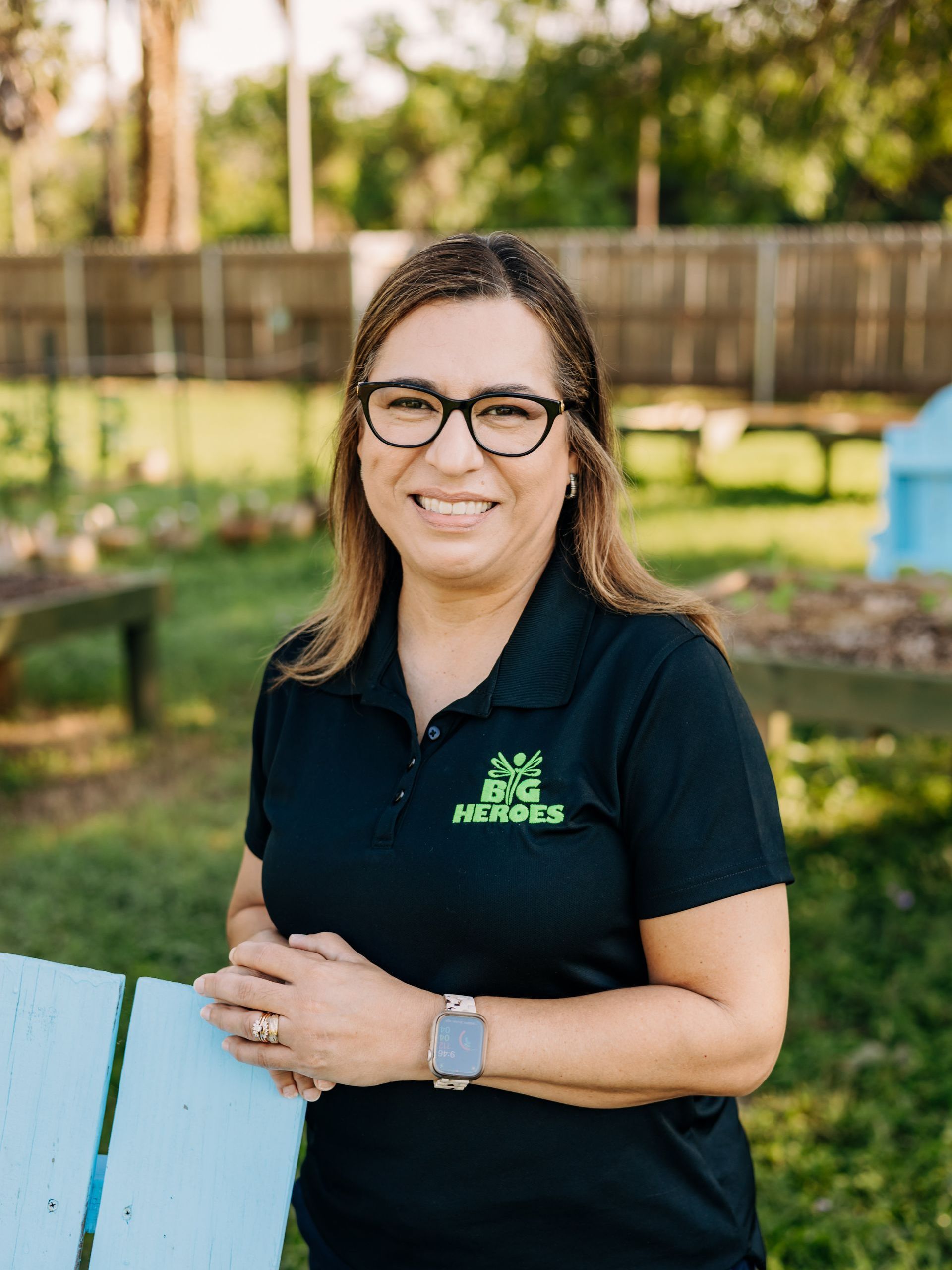 A woman wearing glasses and a black shirt is standing next to a blue bench.