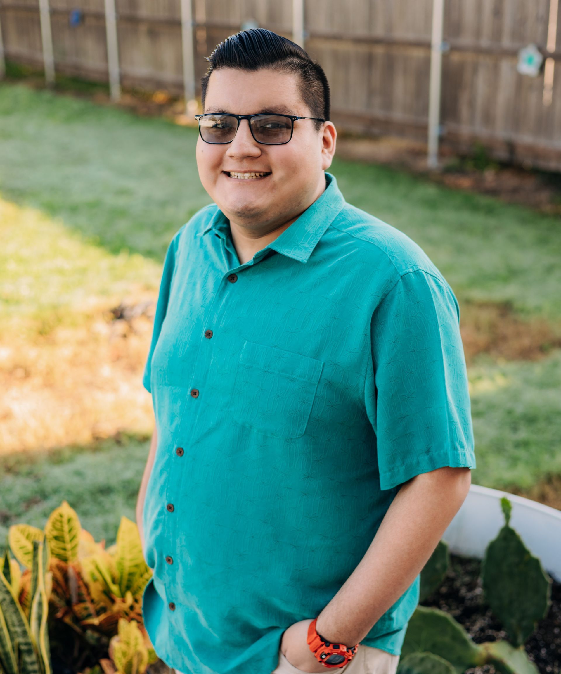 A man wearing sunglasses and a green shirt is standing in a garden.