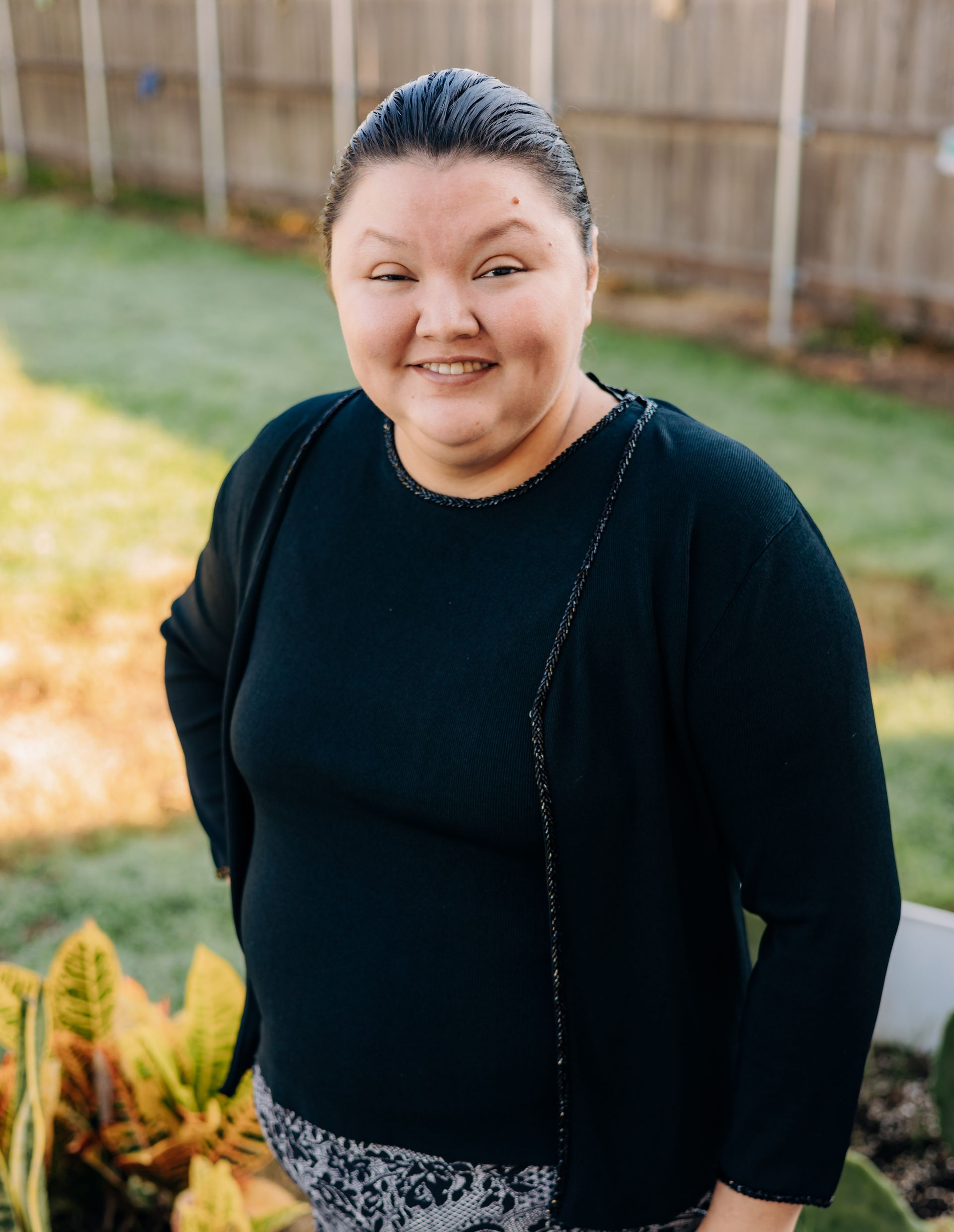 A woman in a black sweater is standing in front of a fence and smiling.