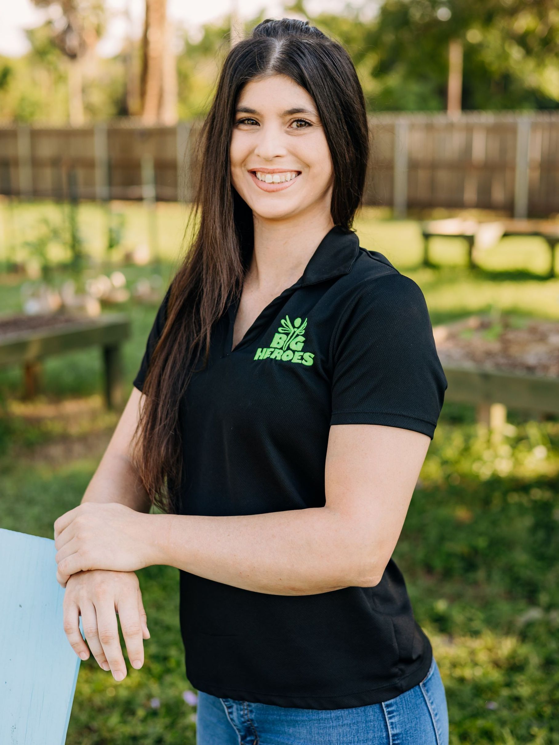 A woman wearing a black polo shirt and blue jeans is standing in a field.