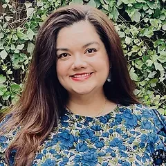 A woman in a blue floral shirt is smiling in front of a bush.