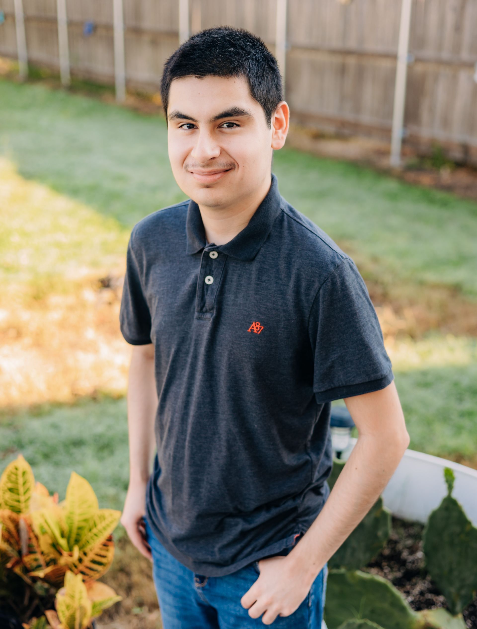 A young man in a black polo shirt is standing in front of a fence.