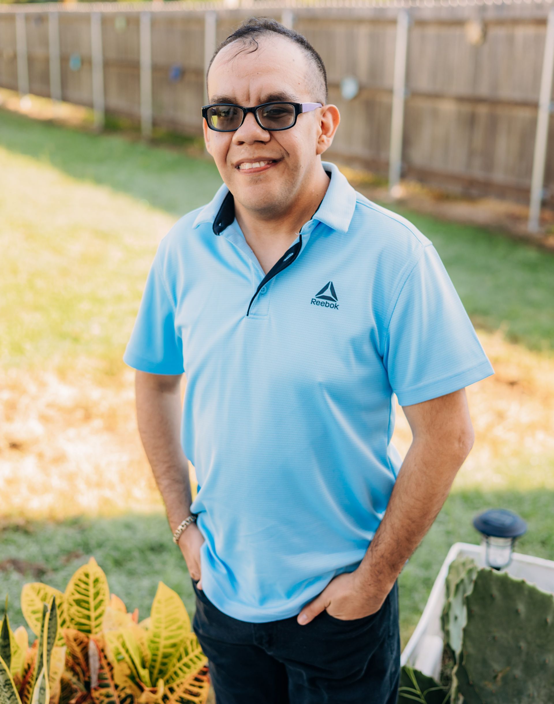 A man wearing glasses and a blue shirt is standing in a yard with his hands in his pockets.
