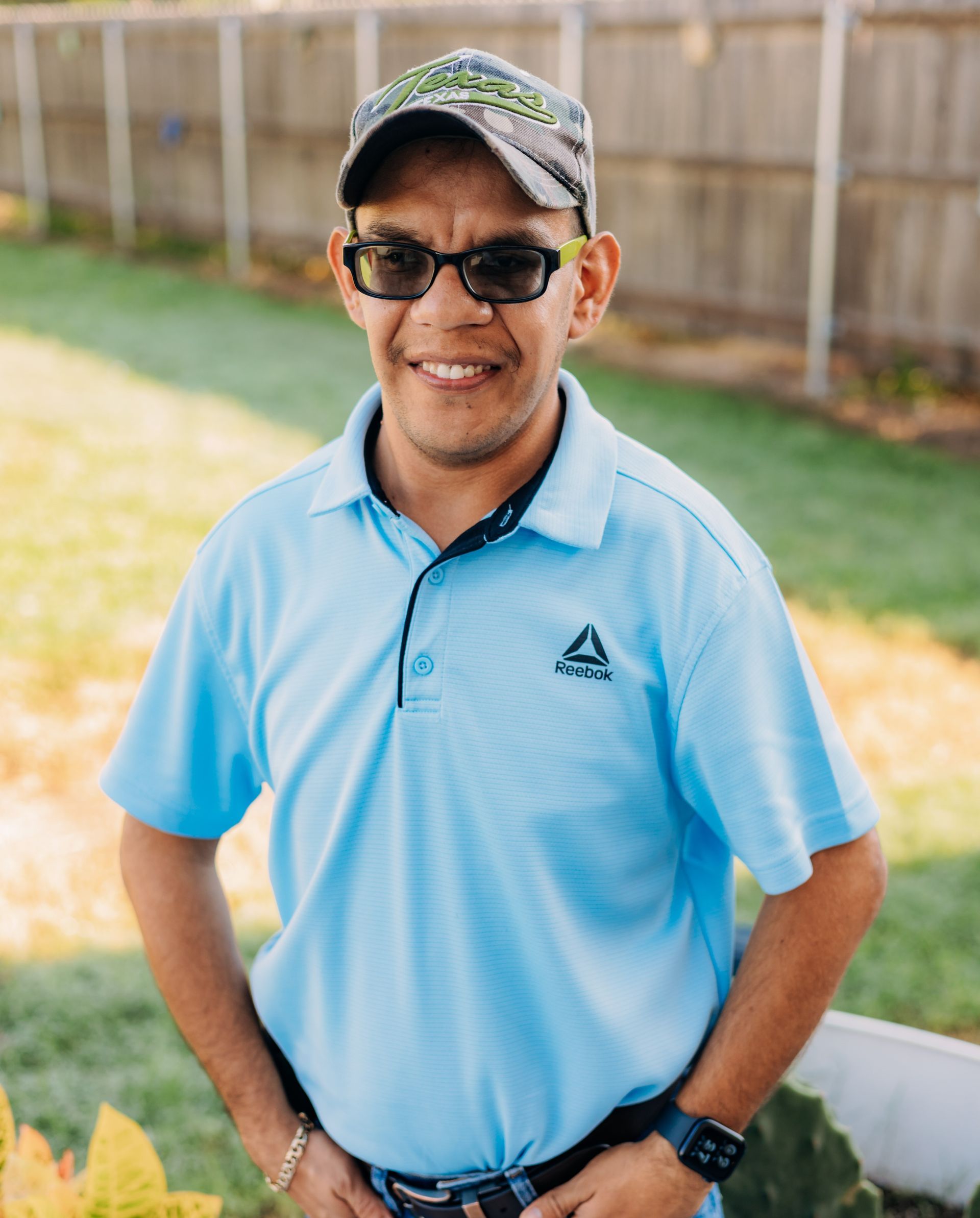 A man wearing a hat and sunglasses is standing in front of a fence.
