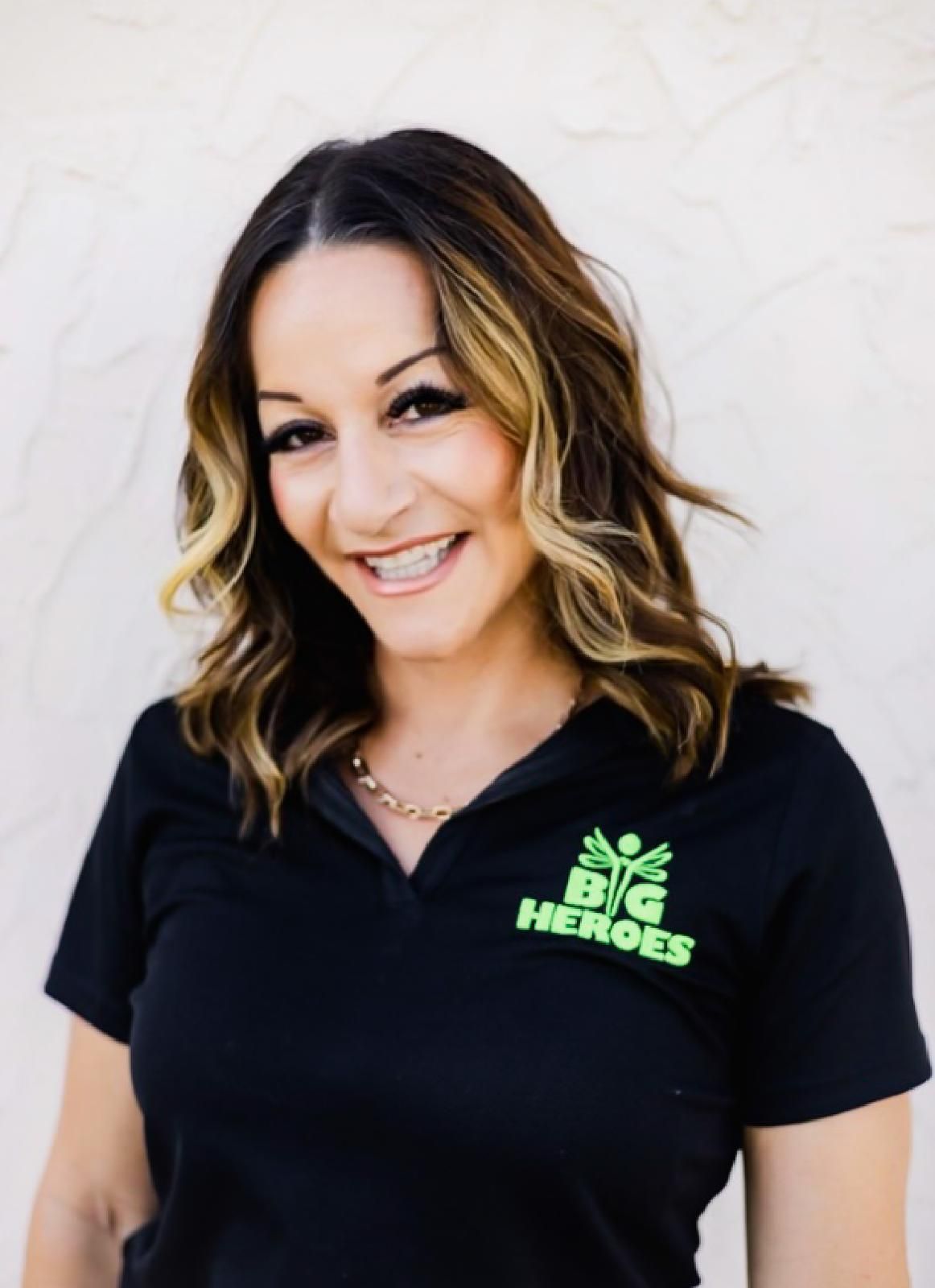 Woman in black shirt with logo smiles in front of a wooden table and green background.