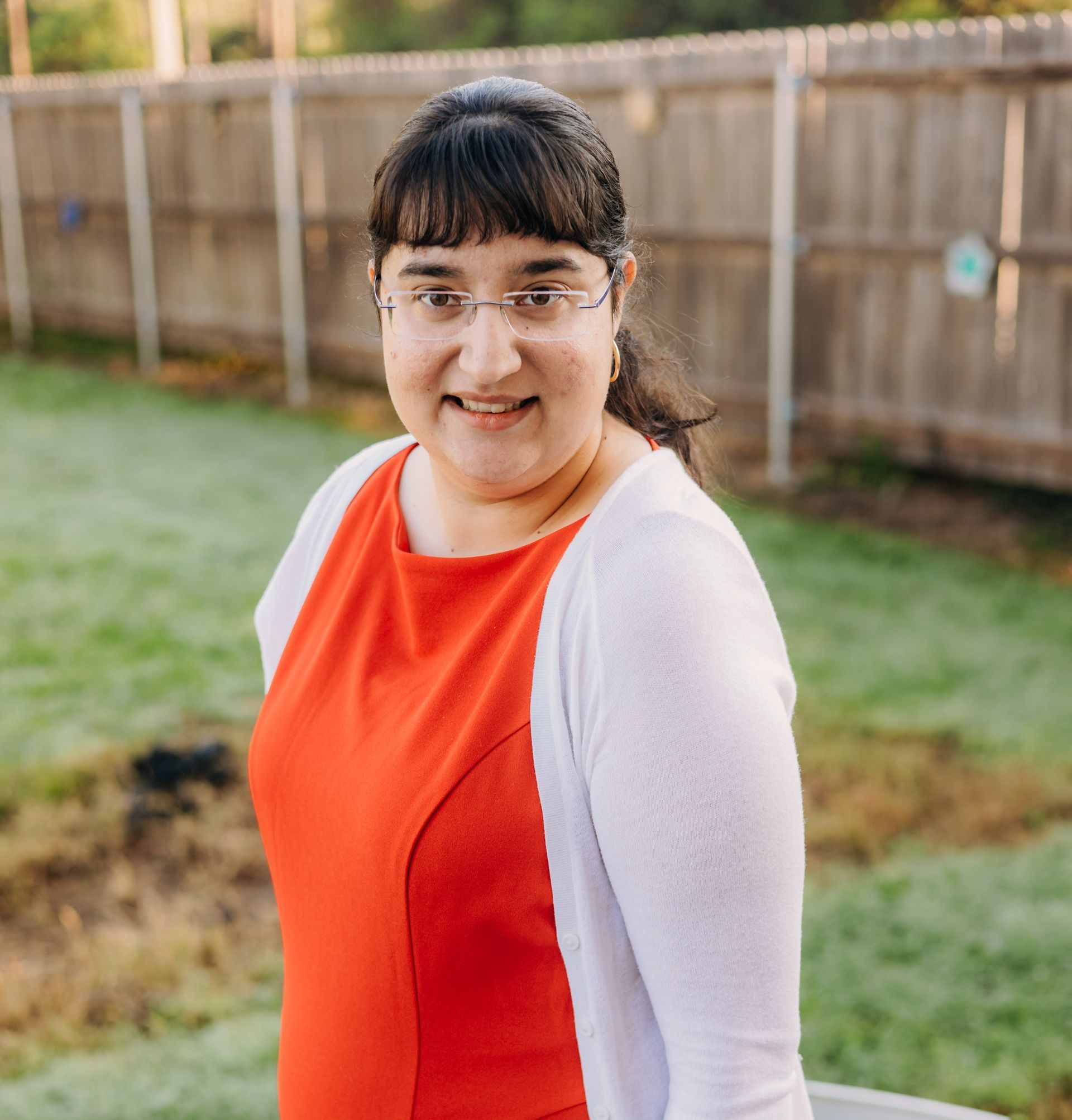 A woman wearing glasses and a red dress is standing in front of a wooden fence.