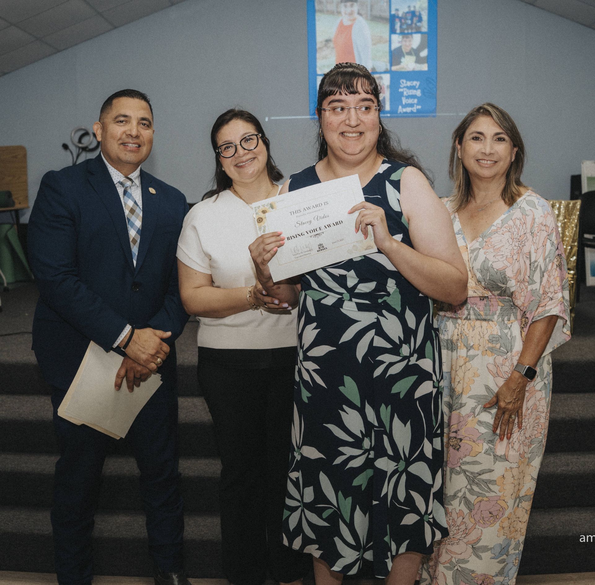 Four people stand indoors; award recipient holds certificate. Others smile. Stage with banner in background.