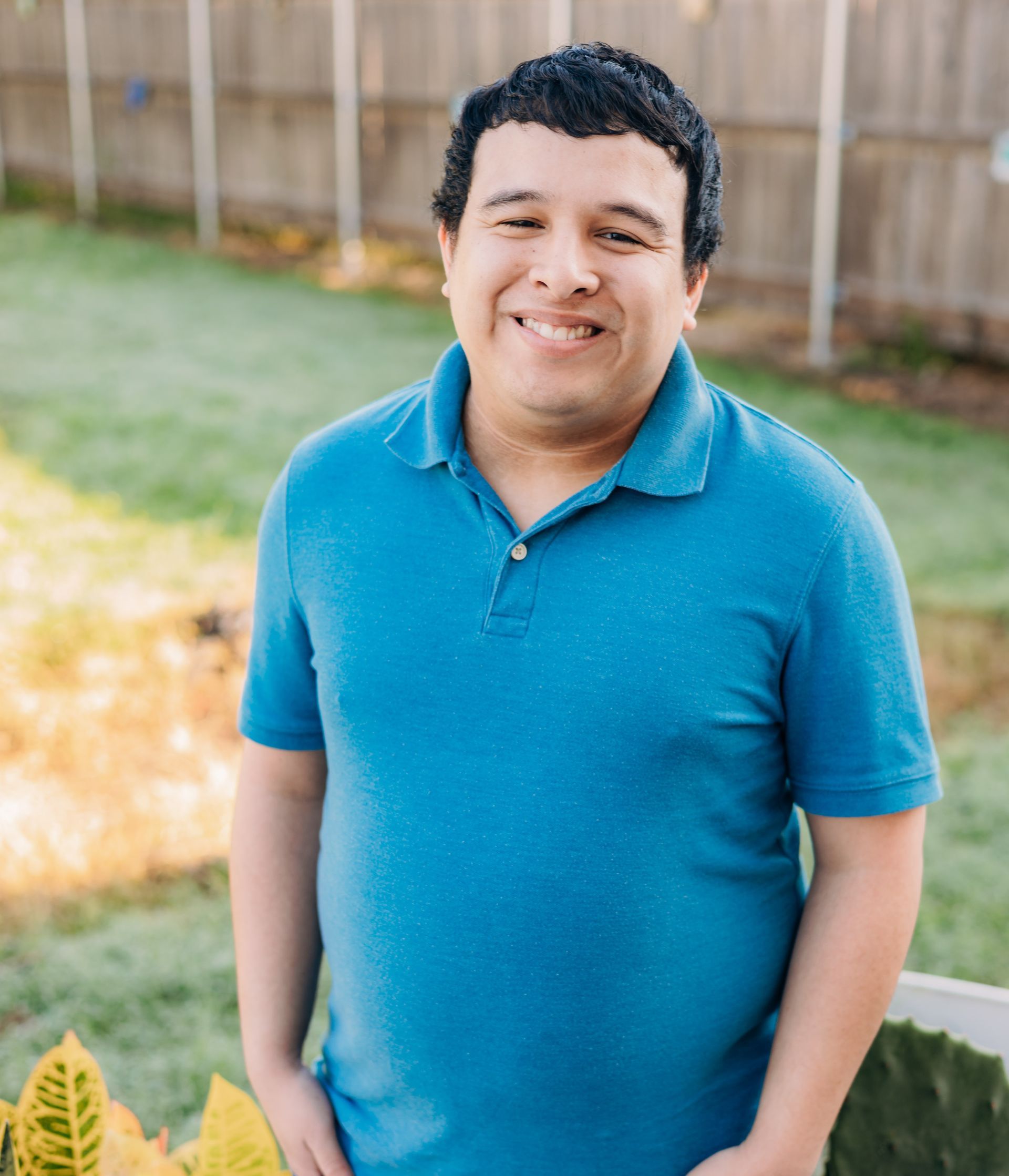A man in a blue shirt is standing in front of a fence and smiling.