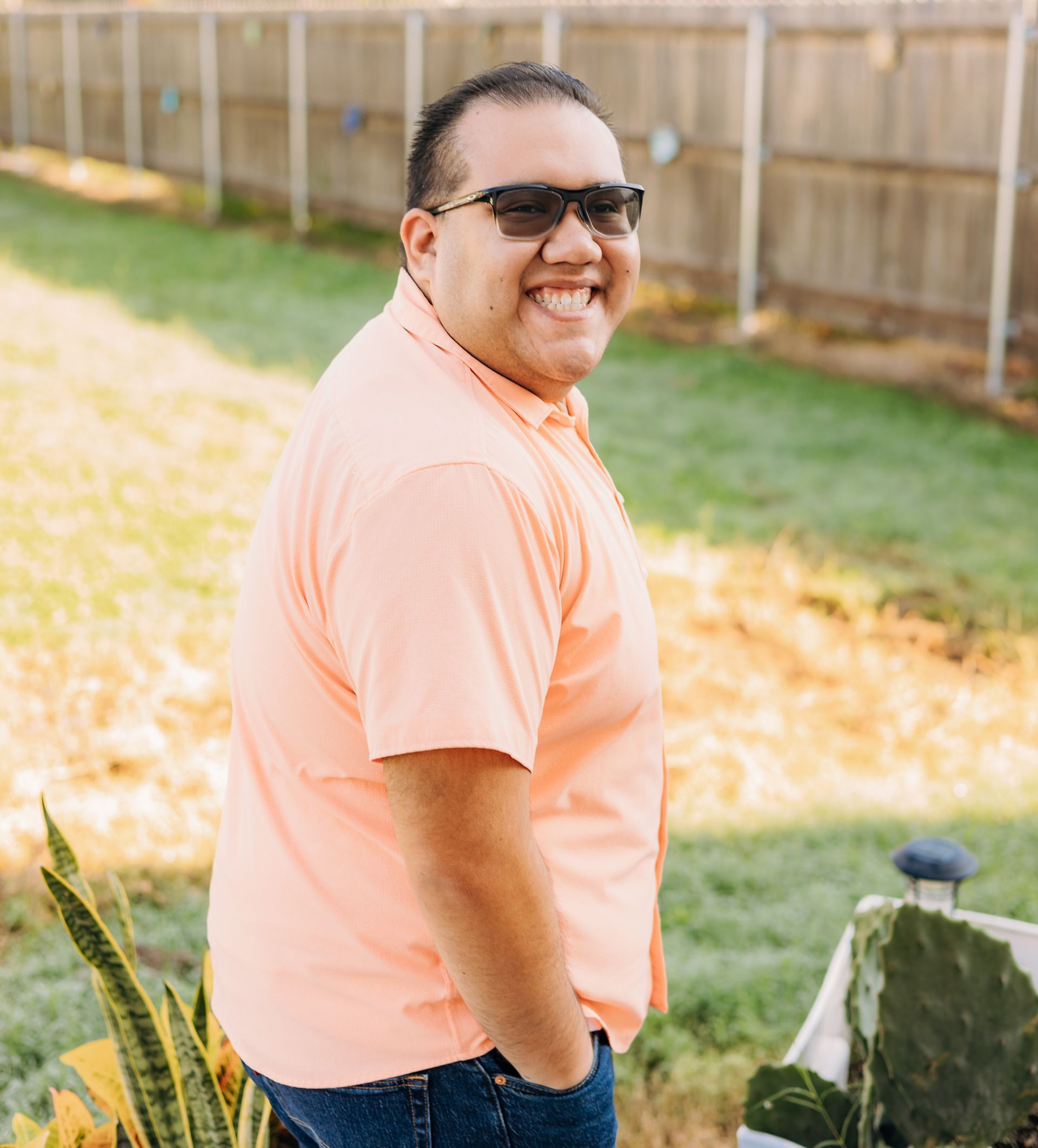 A man wearing sunglasses and a pink shirt is standing in a yard.