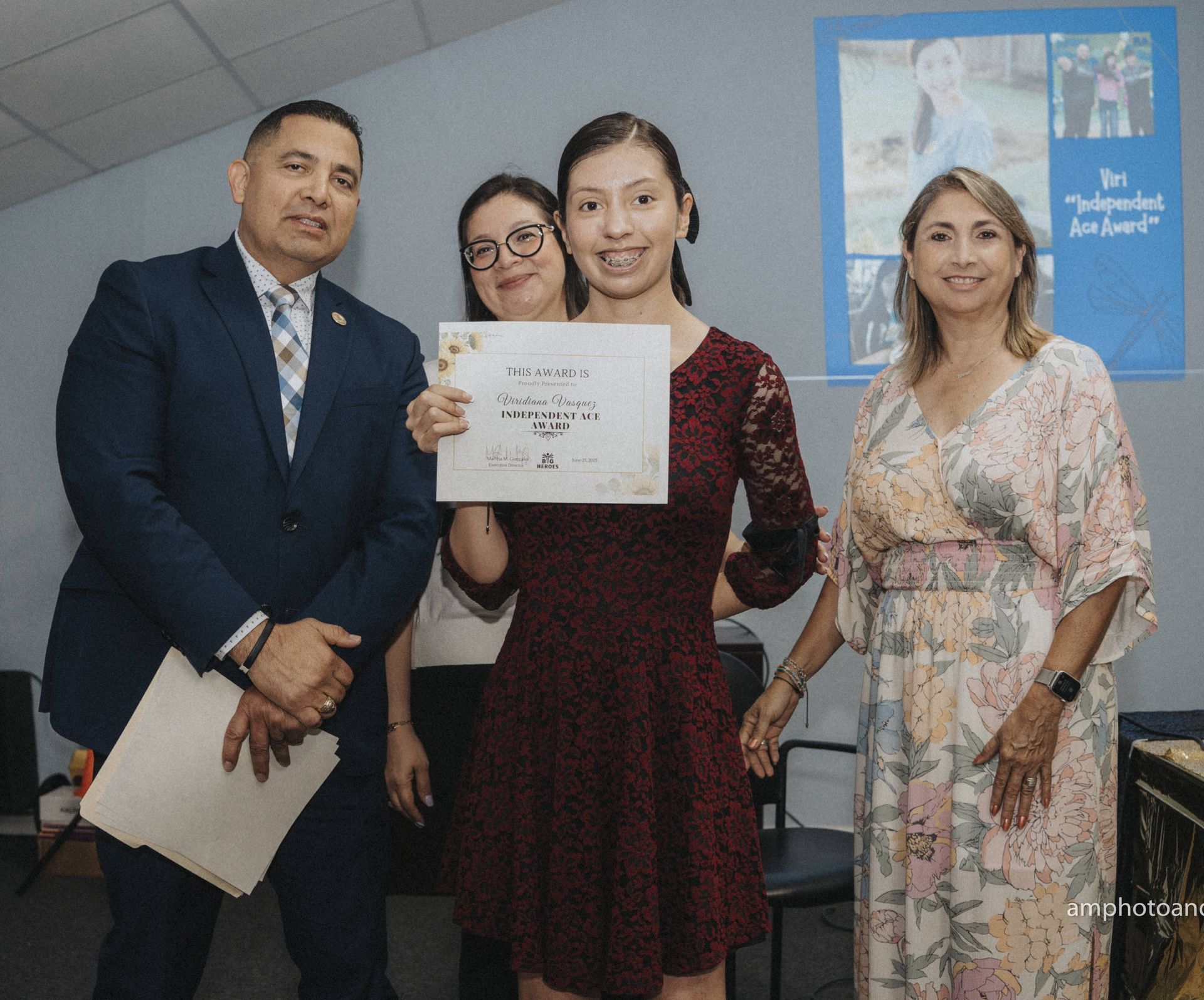 People at an event: a woman in a burgundy dress holding a certificate, flanked by people, indoors.