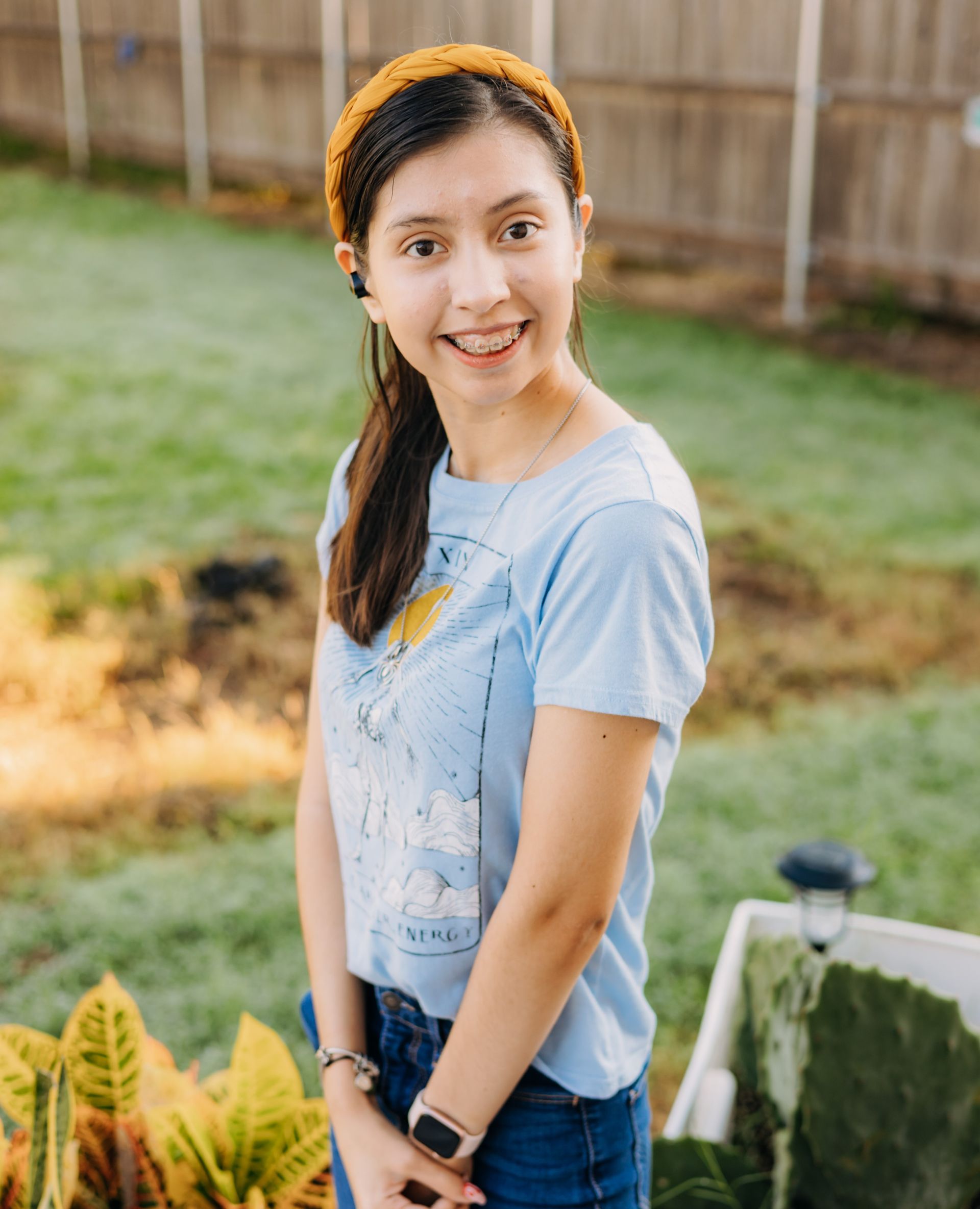 A young woman wearing a blue shirt and a yellow headband is standing in a garden.