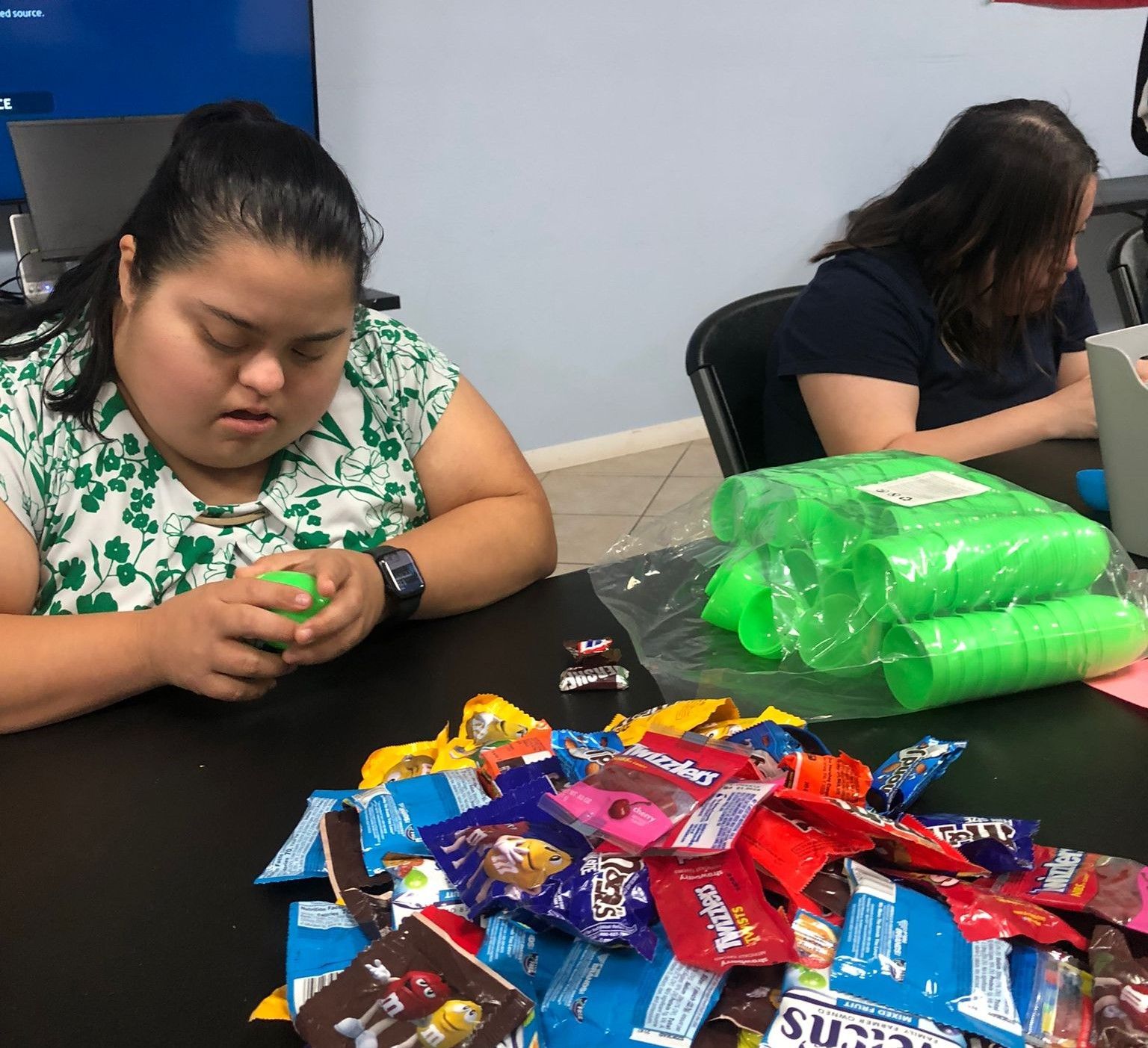 Two women are sitting at a table with a pile of candy and a stack of green cups.
