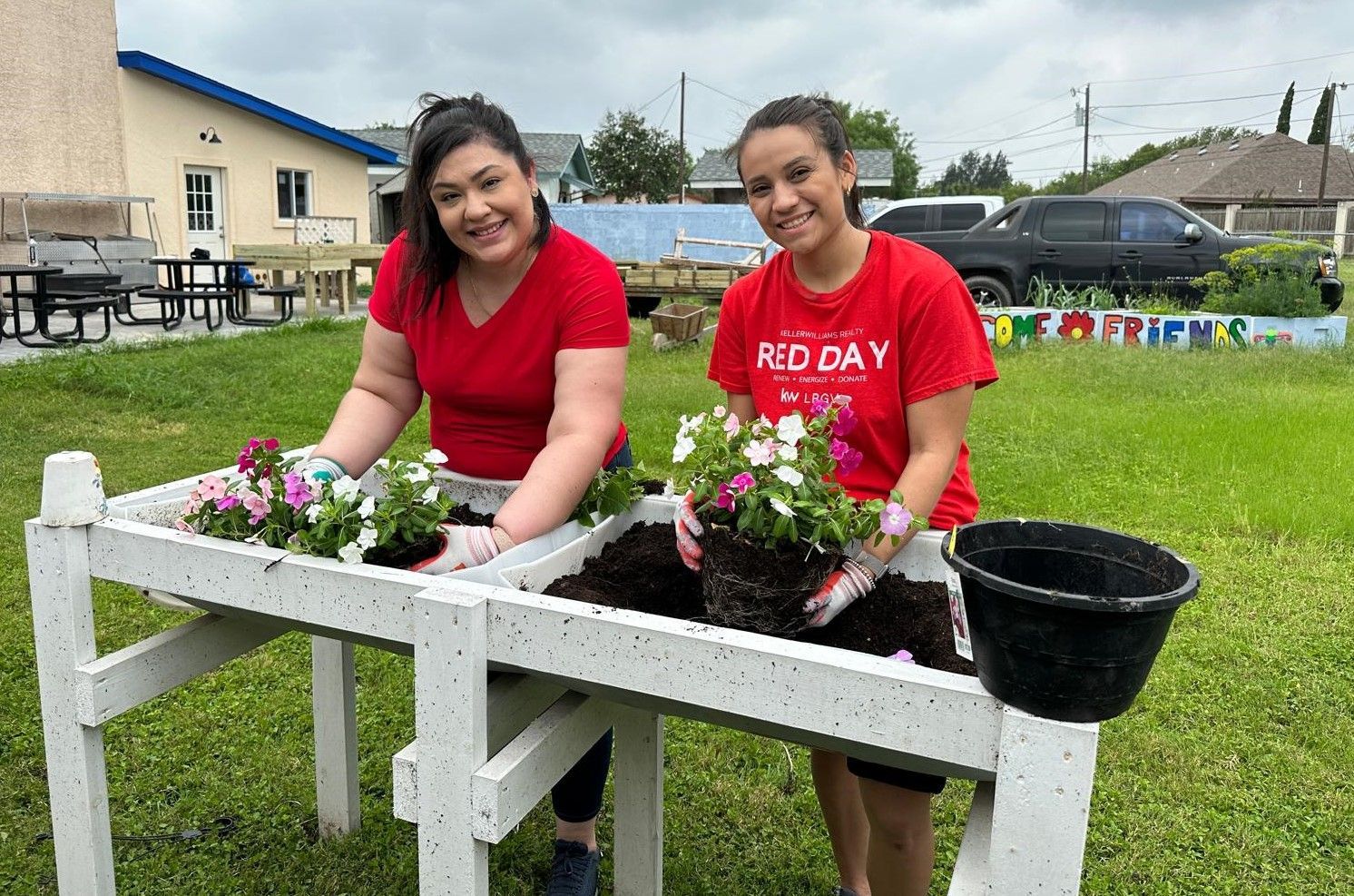 Two women in red shirts are planting flowers in a garden.