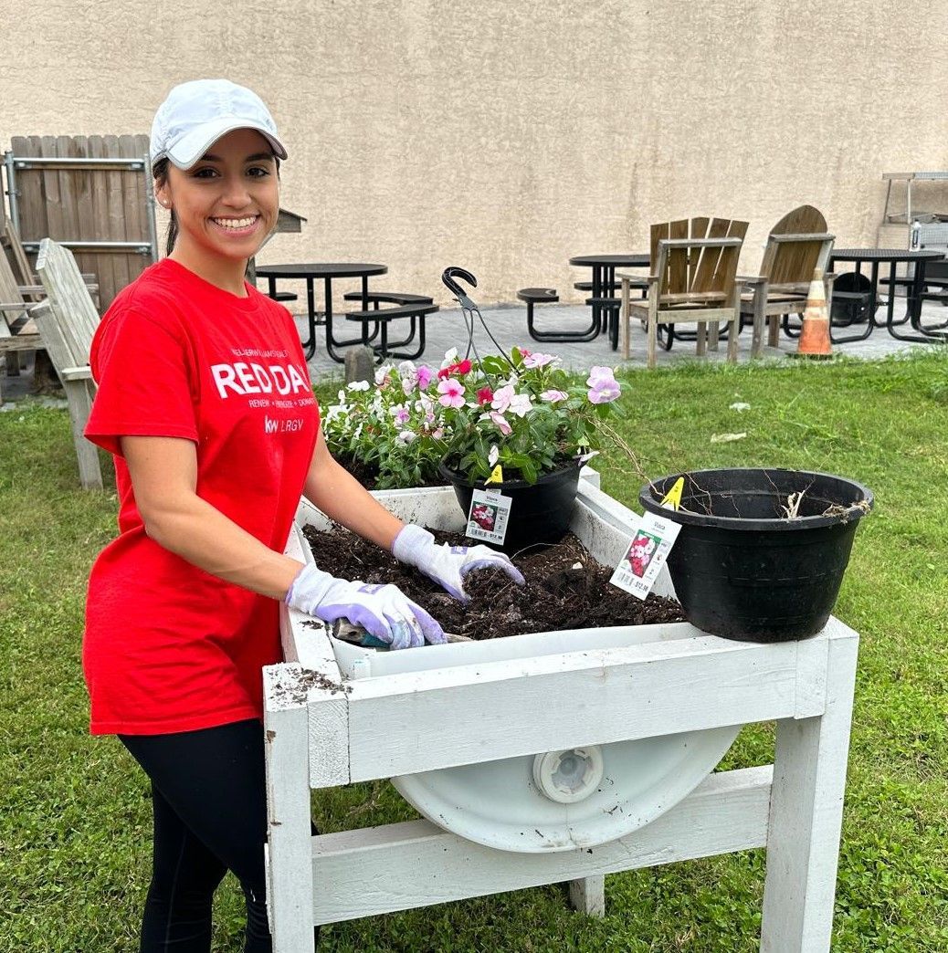 A woman in a red shirt is standing next to a wheelbarrow filled with flowers.