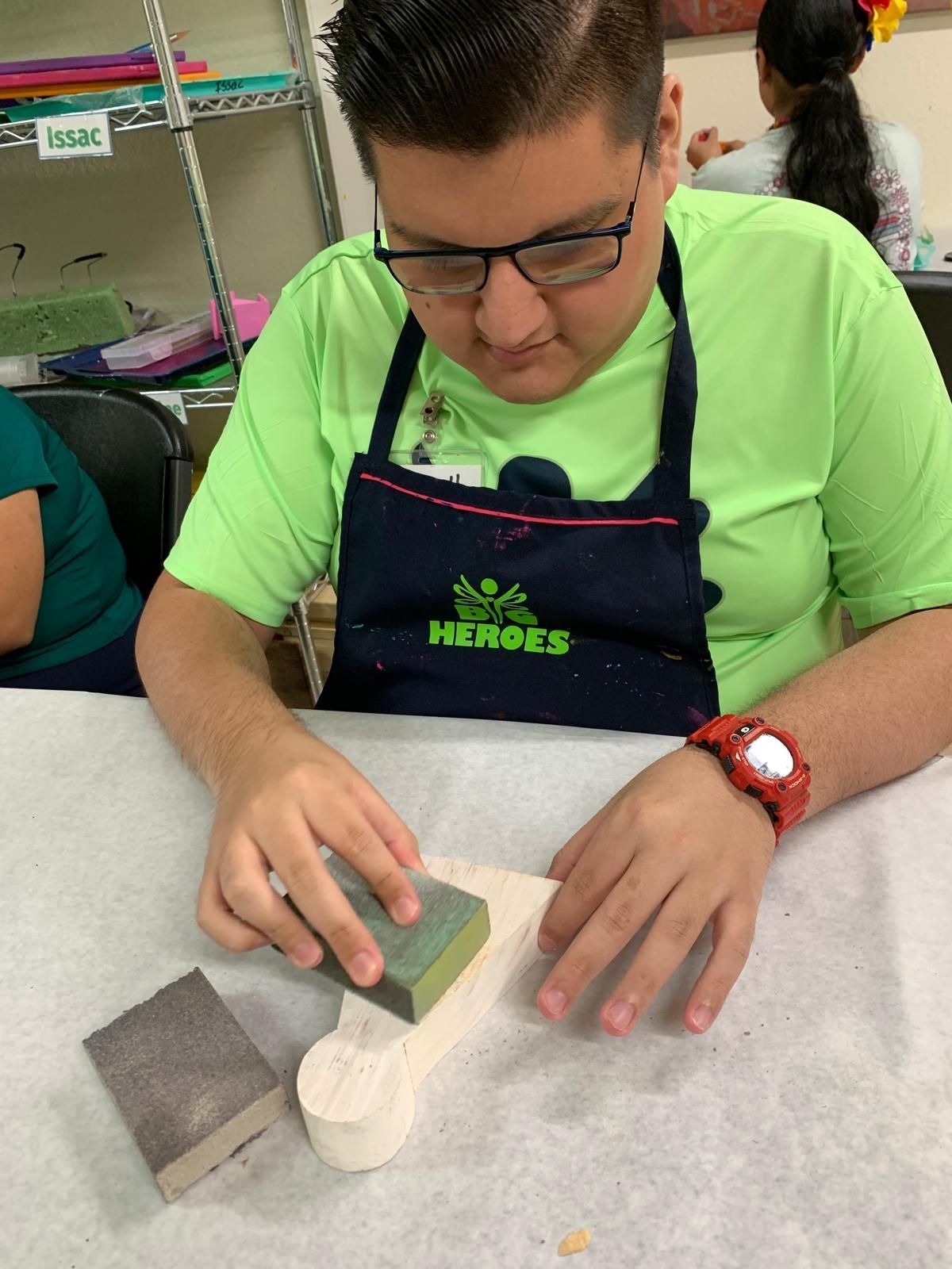 A boy wearing a green shirt and an apron is sanding a piece of wood.