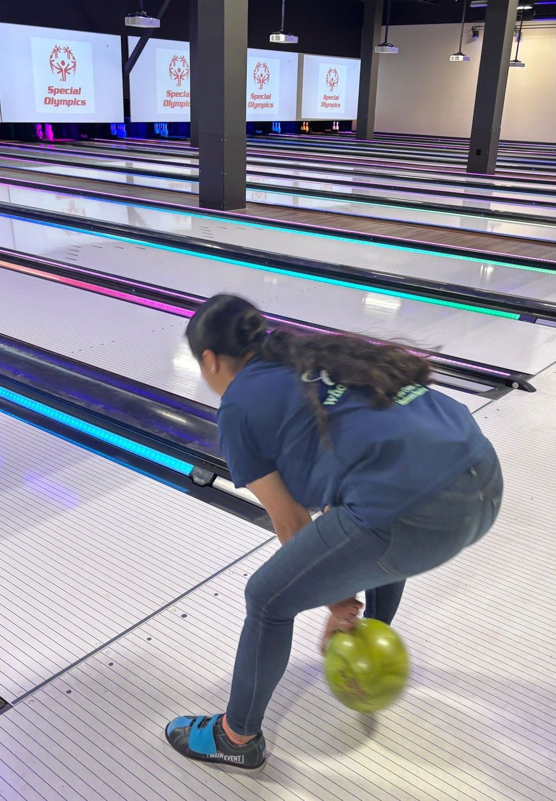 A girl is bending over to throw a bowling ball on a bowling alley.