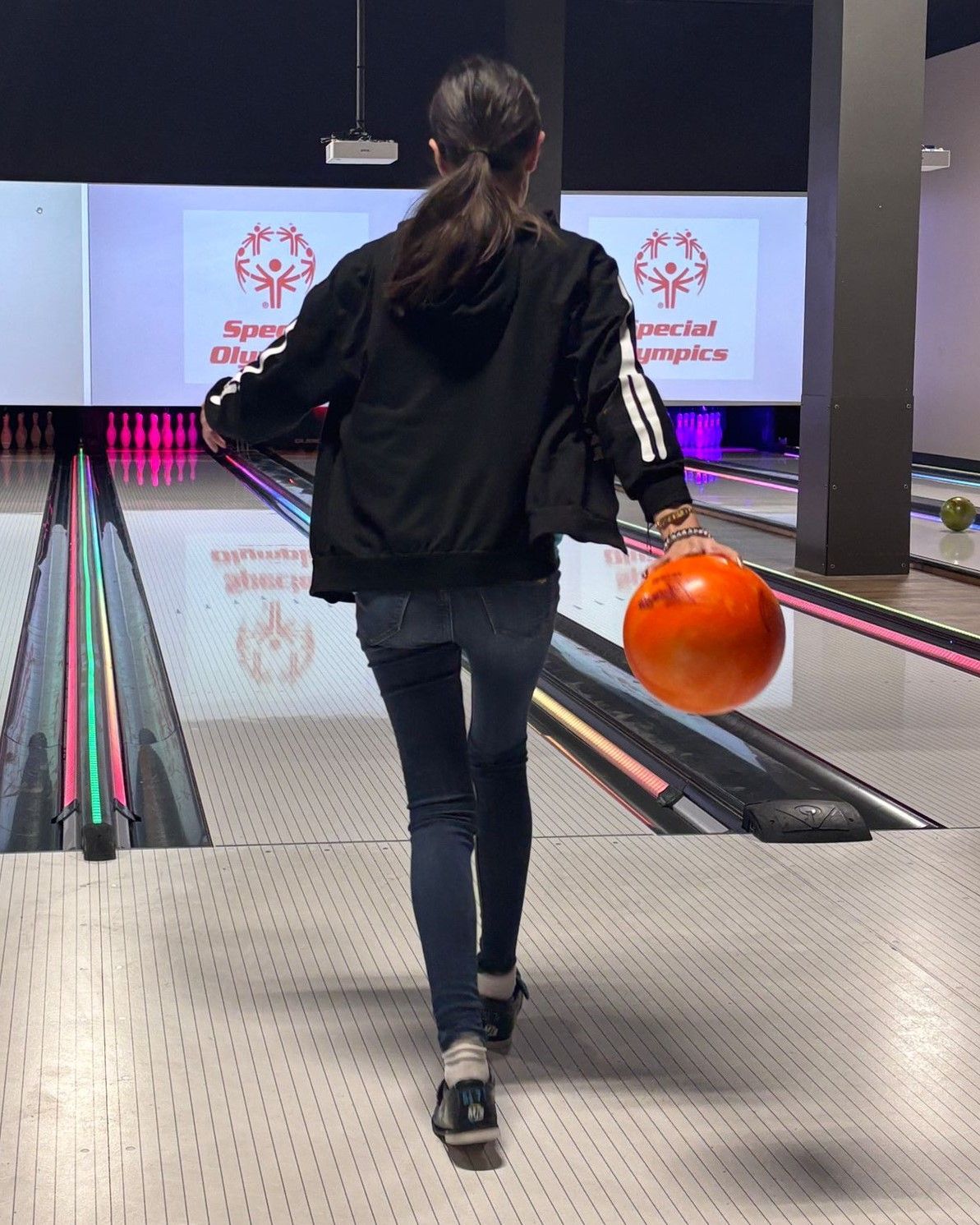 A woman is walking down a bowling alley holding an orange bowling ball.