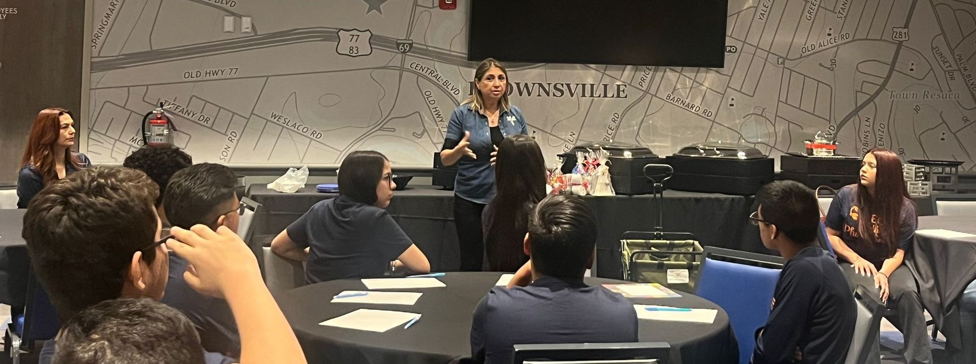 A woman is standing in front of a group of people sitting around a table.