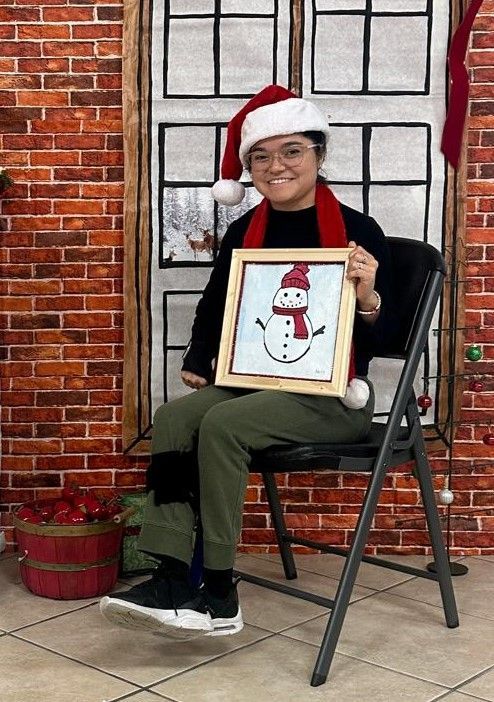 A woman in a santa hat is sitting in a chair holding a picture of a snowman.