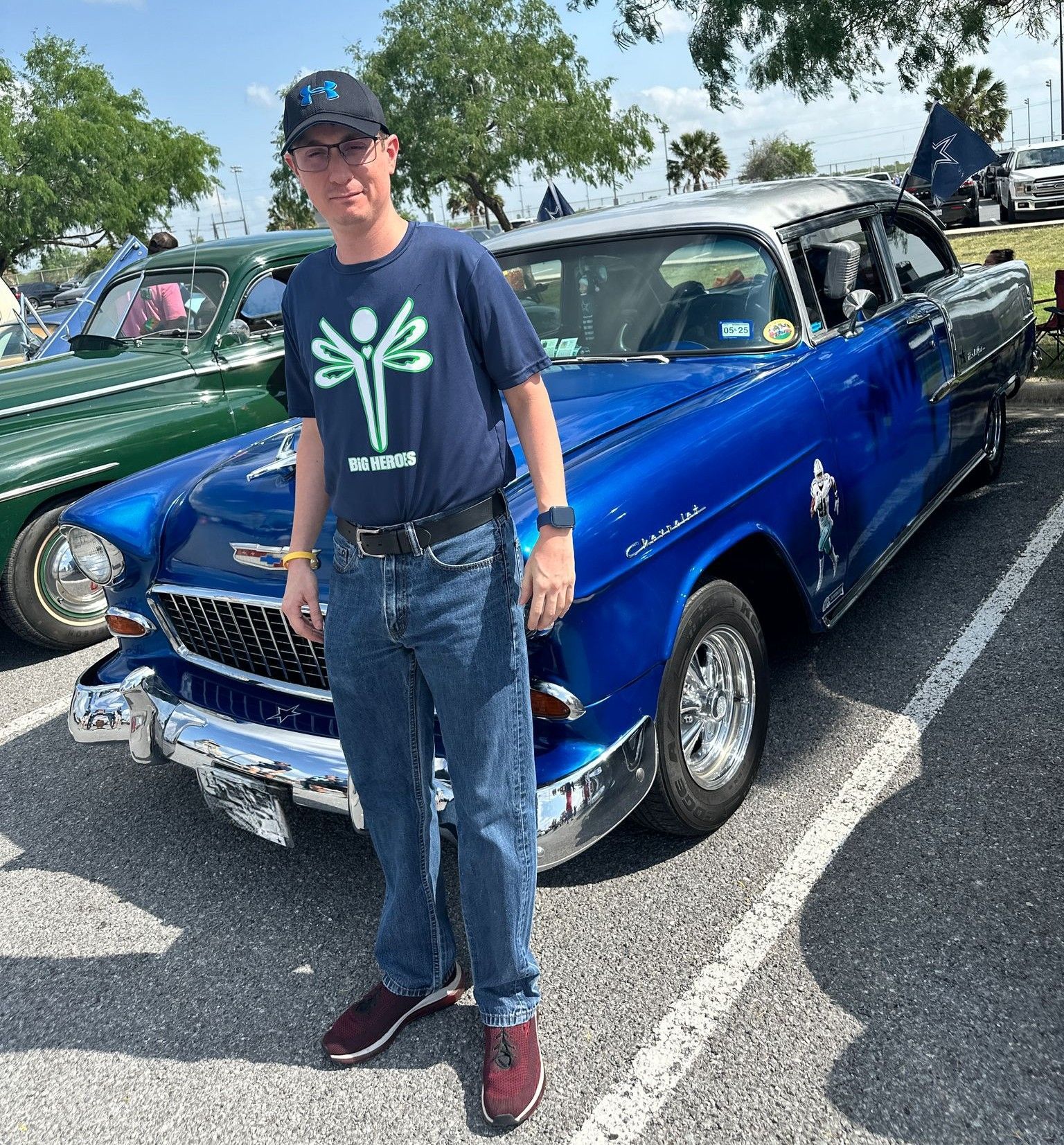 A man is standing in front of a blue car in a parking lot.