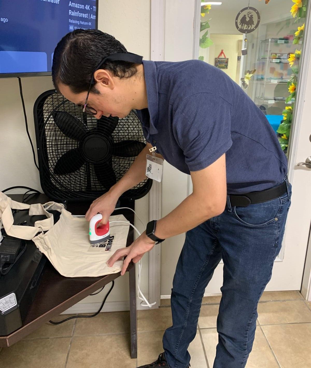 A man is ironing a tote bag on a table in front of a fan.