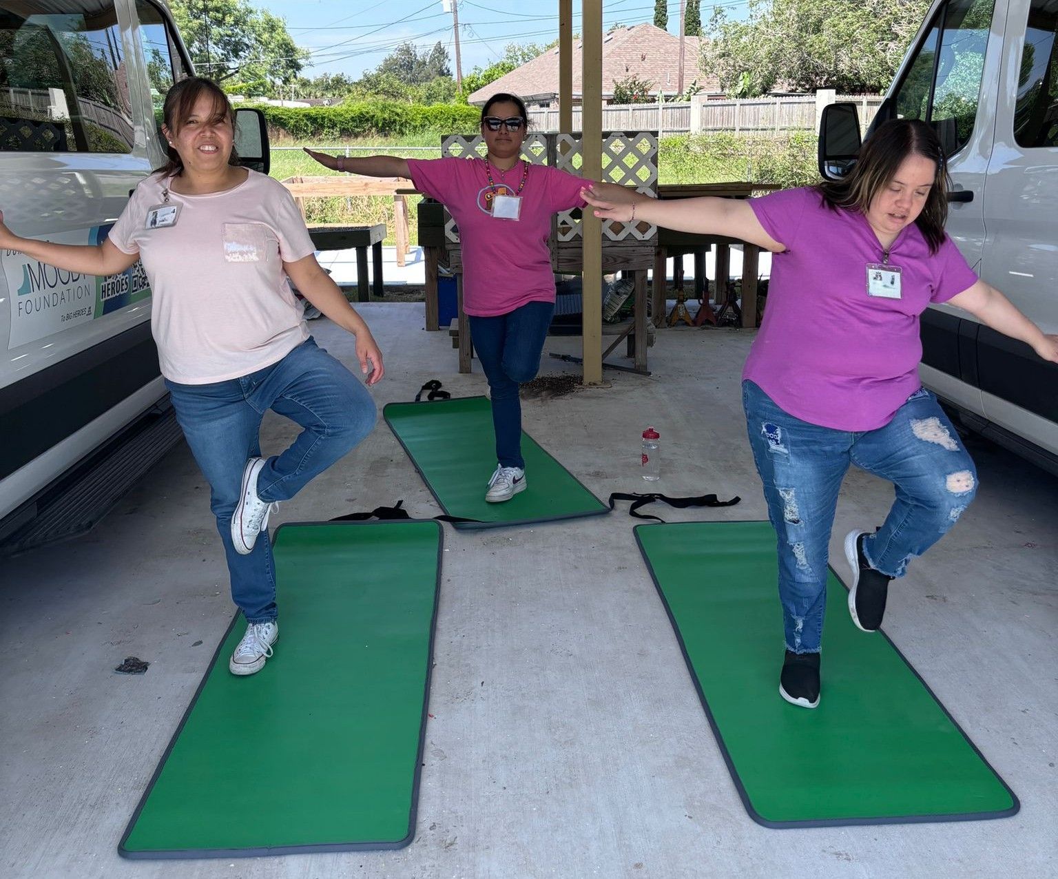 Three women in casual clothes doing a balancing yoga pose on green mats outdoors.