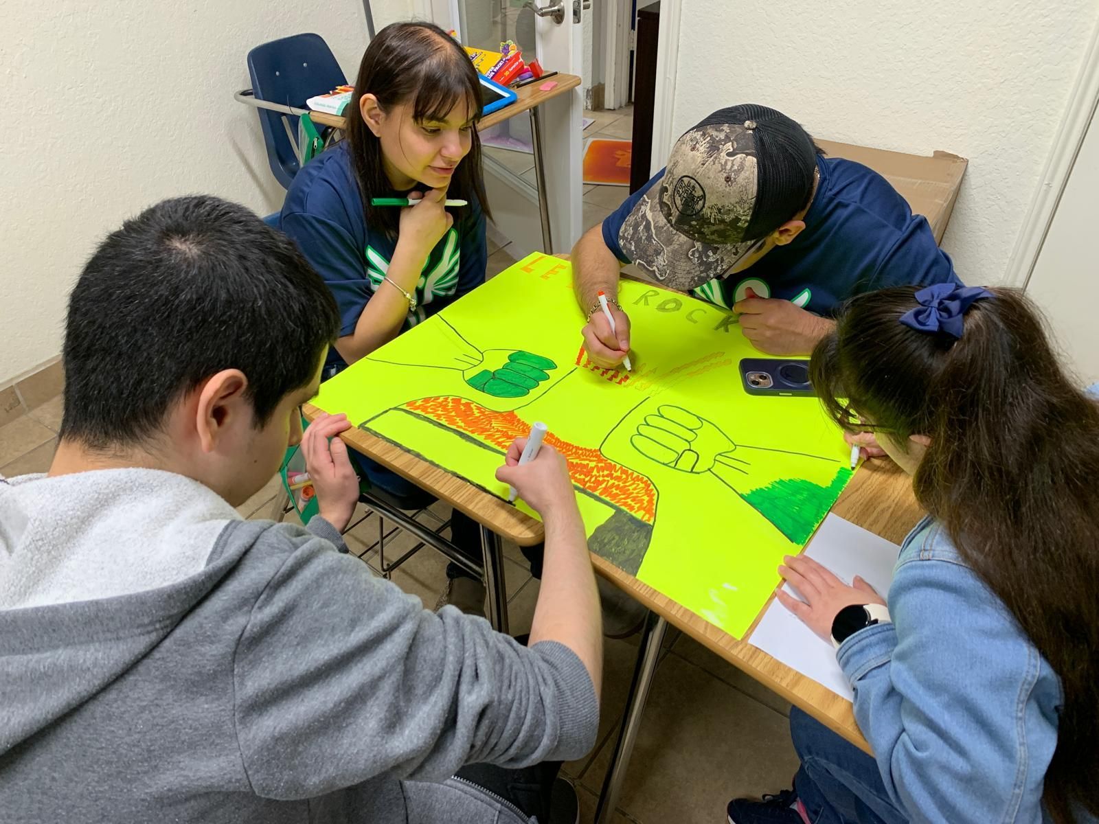 Four people drawing on a large, yellow poster at a desk, likely collaborating on a project.