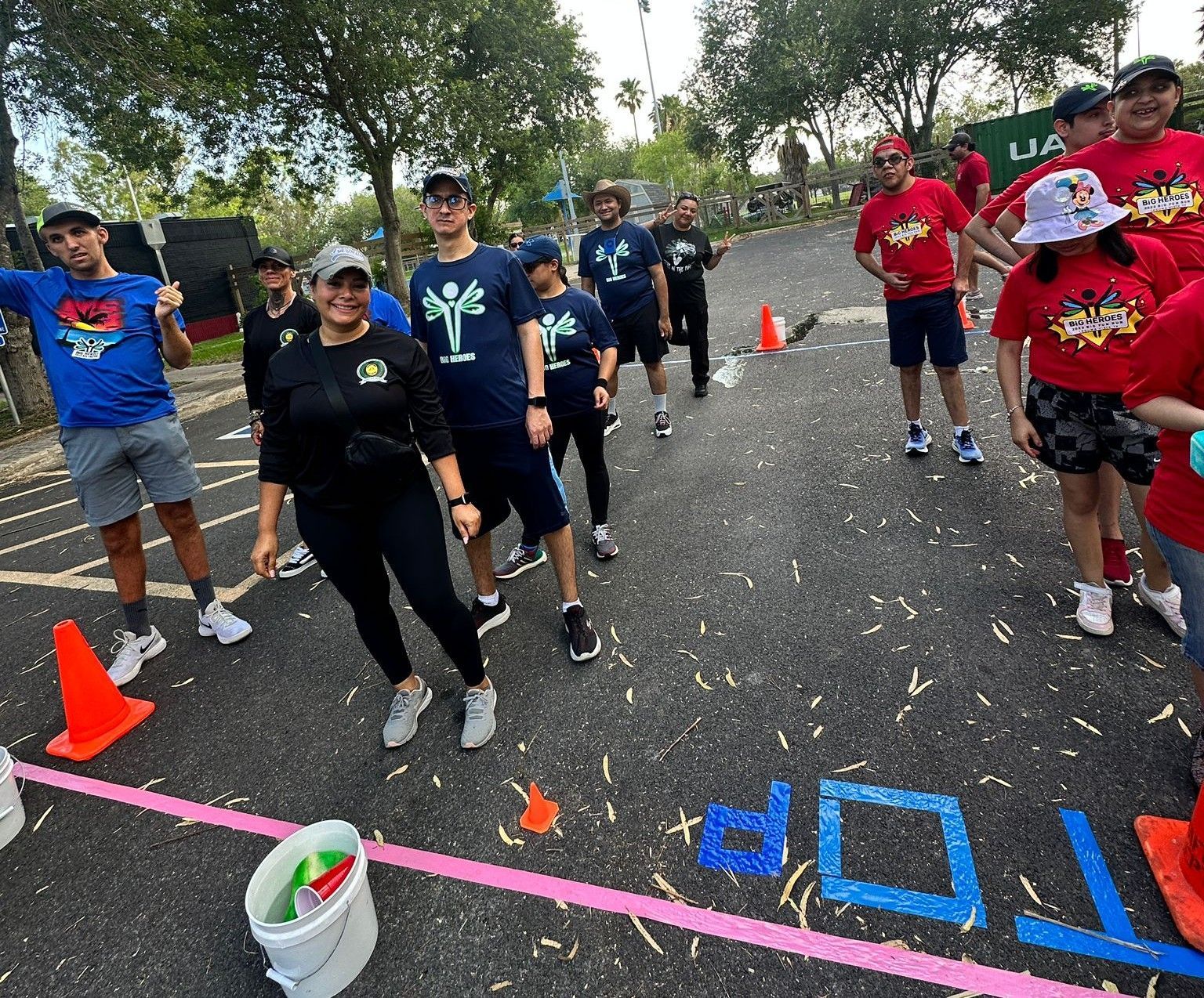 Group of people at a race start line, some in red shirts, some in blue, smiling. Outdoor setting with orange cones.