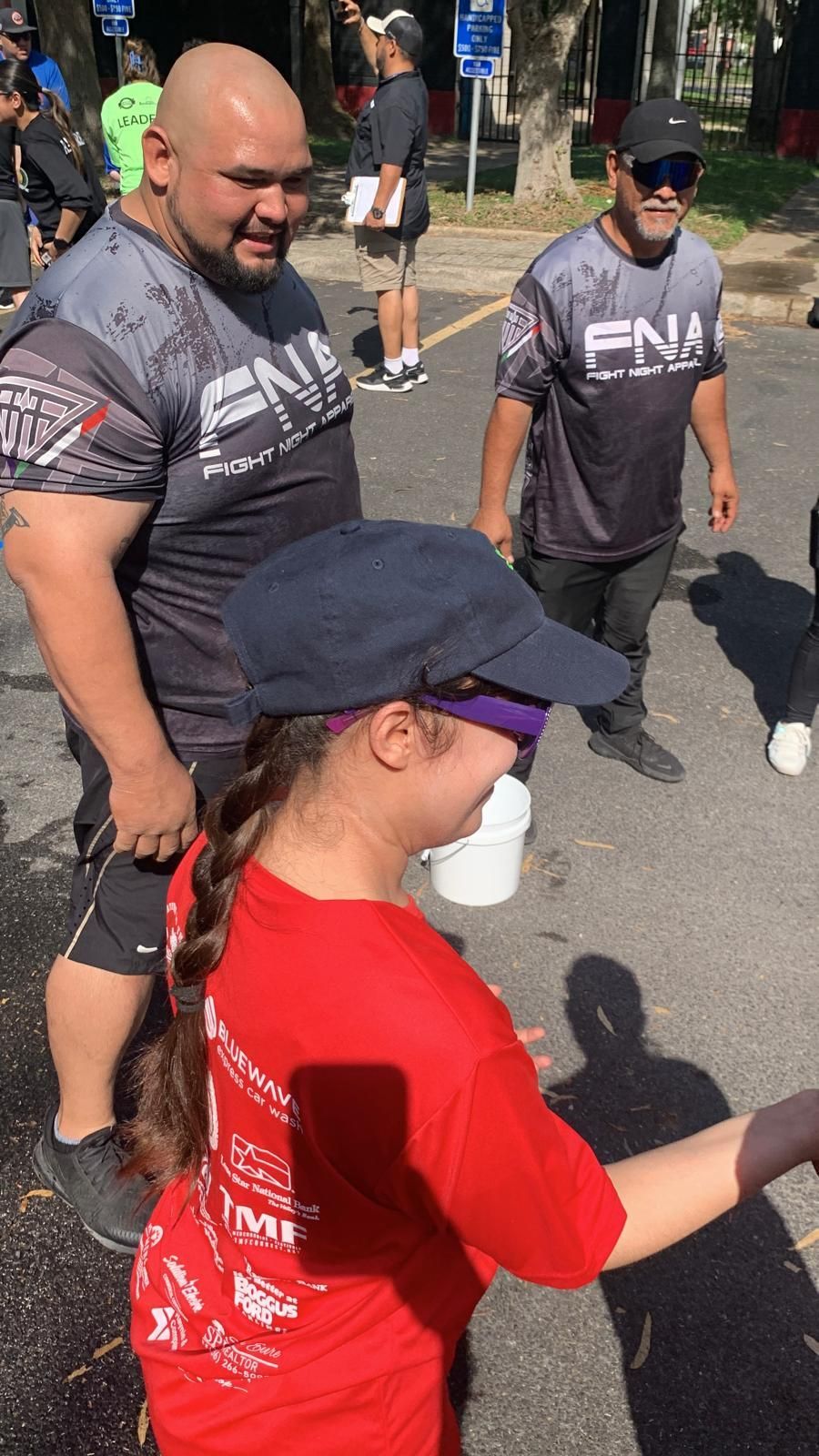 A girl in a red shirt interacts with two men wearing athletic shirts outdoors. Sunny day.