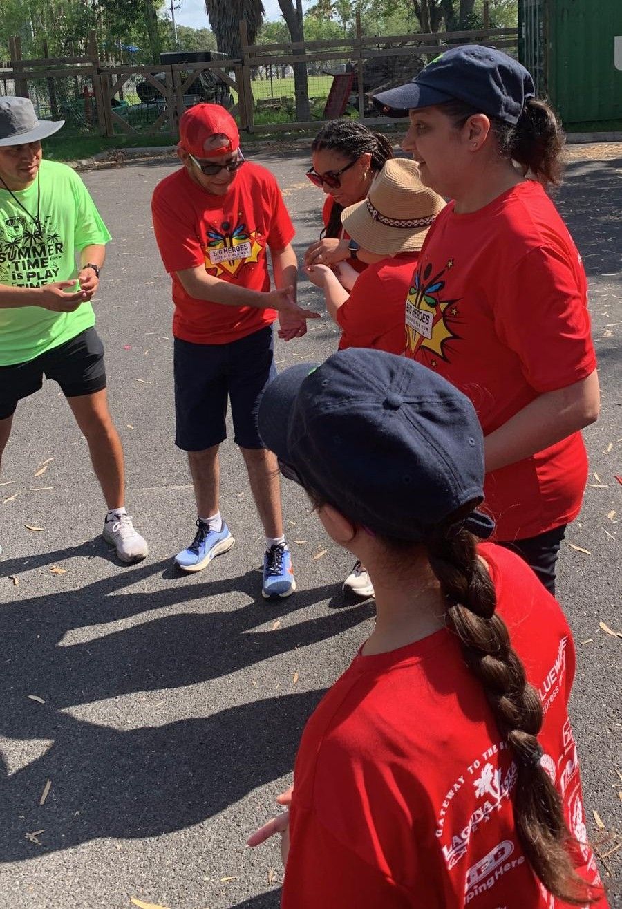 Group of people in red shirts outdoors, one in a hat with a braid. Sunlight casts shadows.