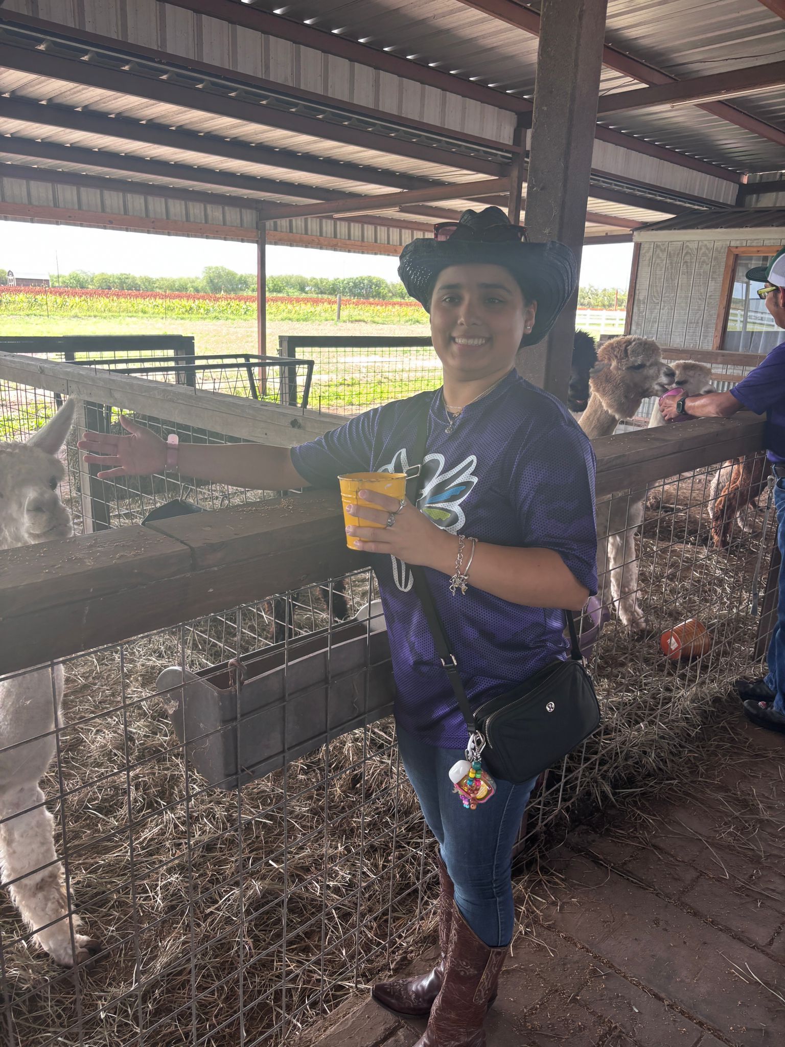 Woman in hat, purple shirt, and jeans smiles, holding a cup at a farm, by a fence.