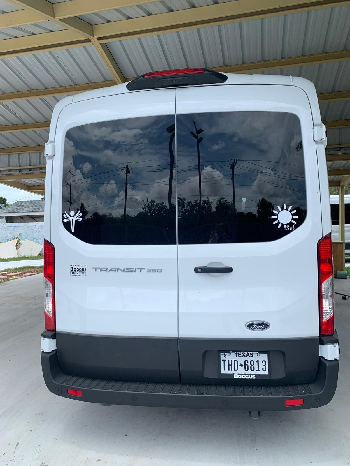 White Ford Transit van, parked under a carport, rear view with stickers on the tinted back window.